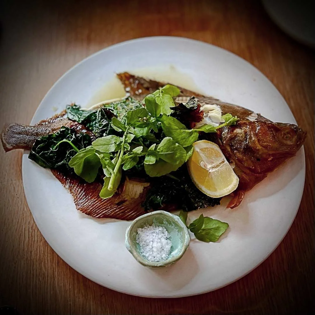 Pan-fried fish from Iluka restaurant in Copenhagen, served on a white plate with green leaves, a lemon wedge, coarse salt in a small bowl and a wooden table background.