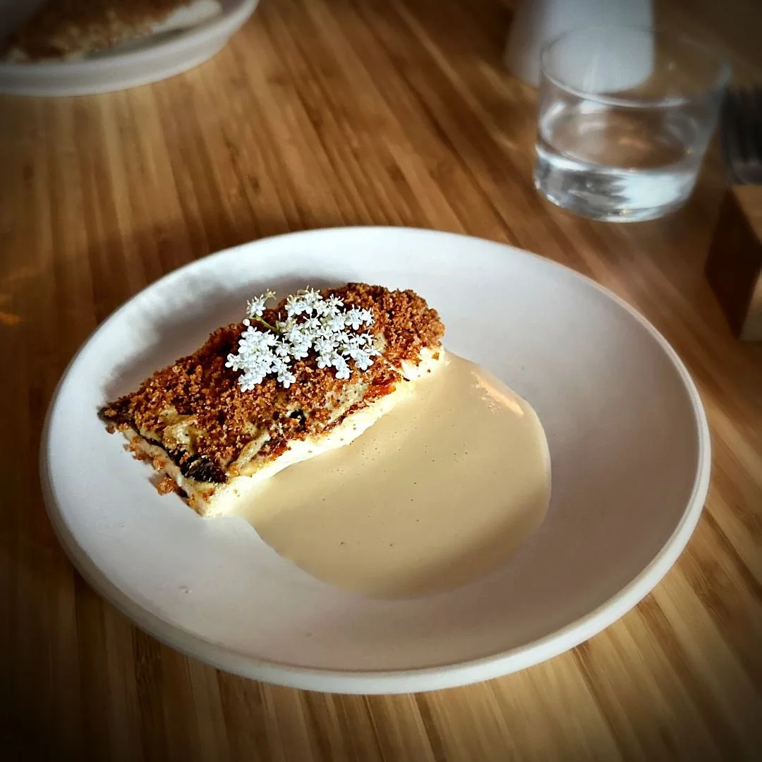 A fillet of fish with crispy crust, served on a light sauce and topped with white blossoms, presented on a white plate at Ja Restaurant in Amsterdam.