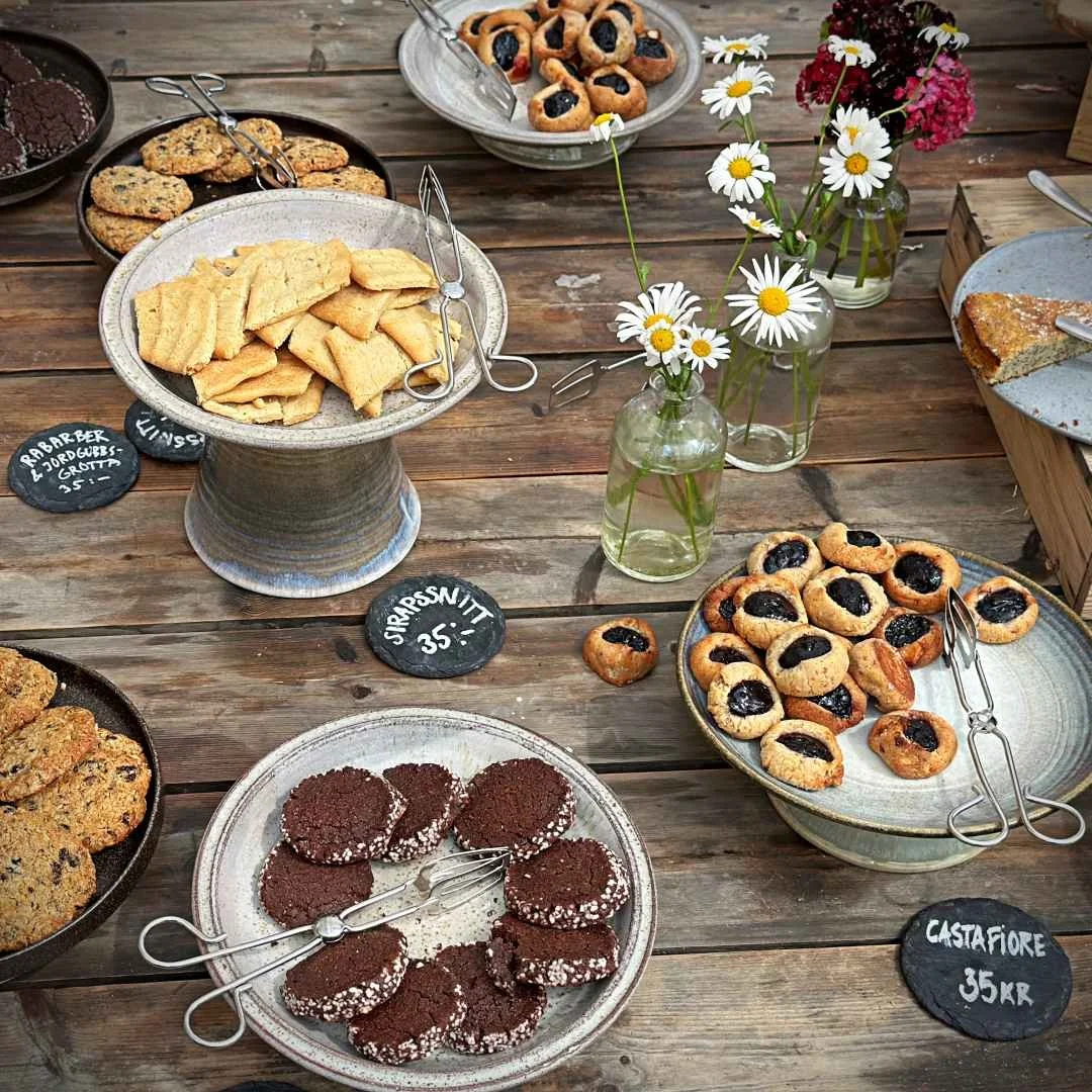 A selection of various cookies on plates and cake stands arranged on a rustic wooden table. Small serving tongs, slate price tags, and small glass bottles with daisies are placed next to them.