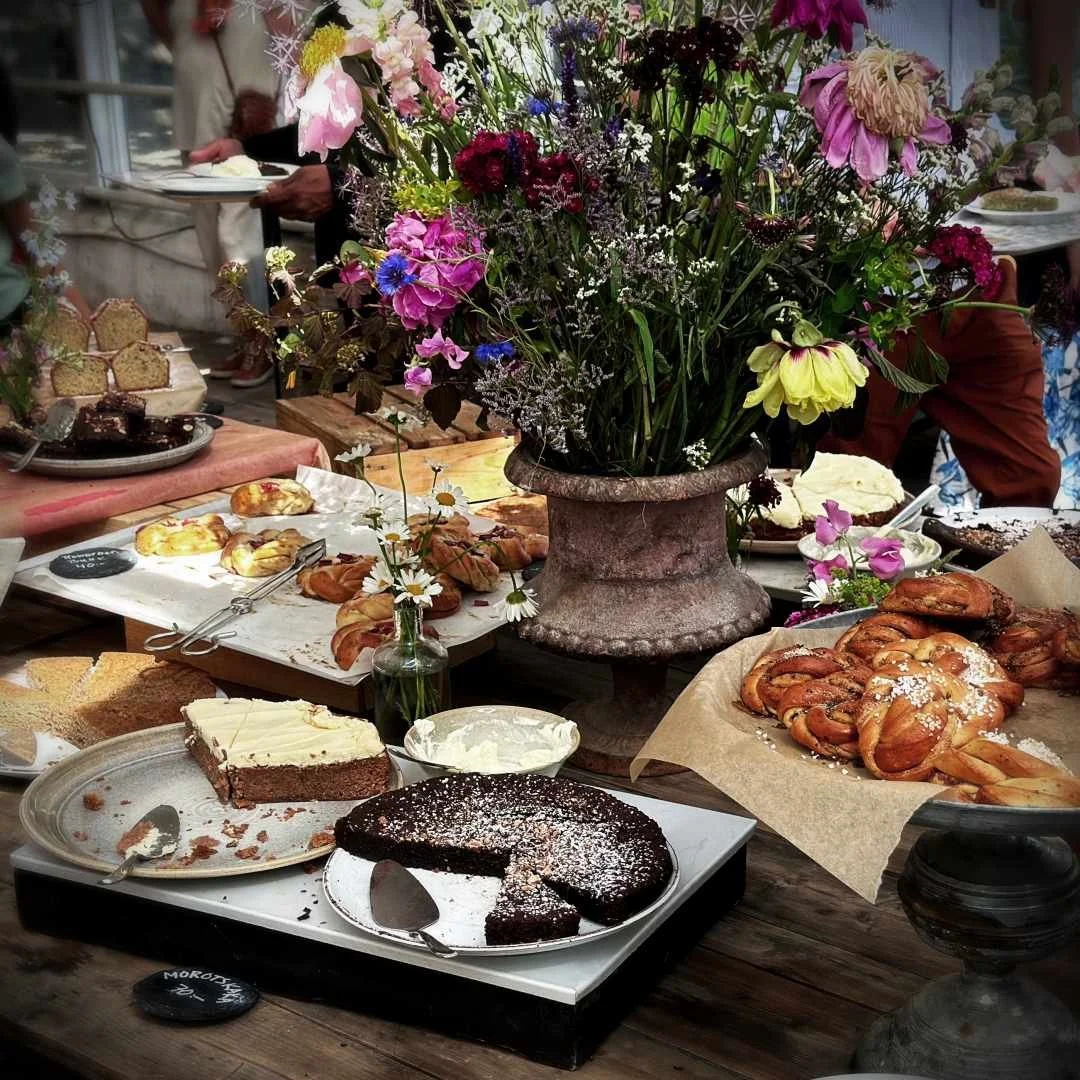 A rich cake buffet on wooden tables featuring cinnamon buns, chocolate cake, carrot cake, and pastries. A large stone vase filled with a colorful bouquet of summer flowers stands in the center.