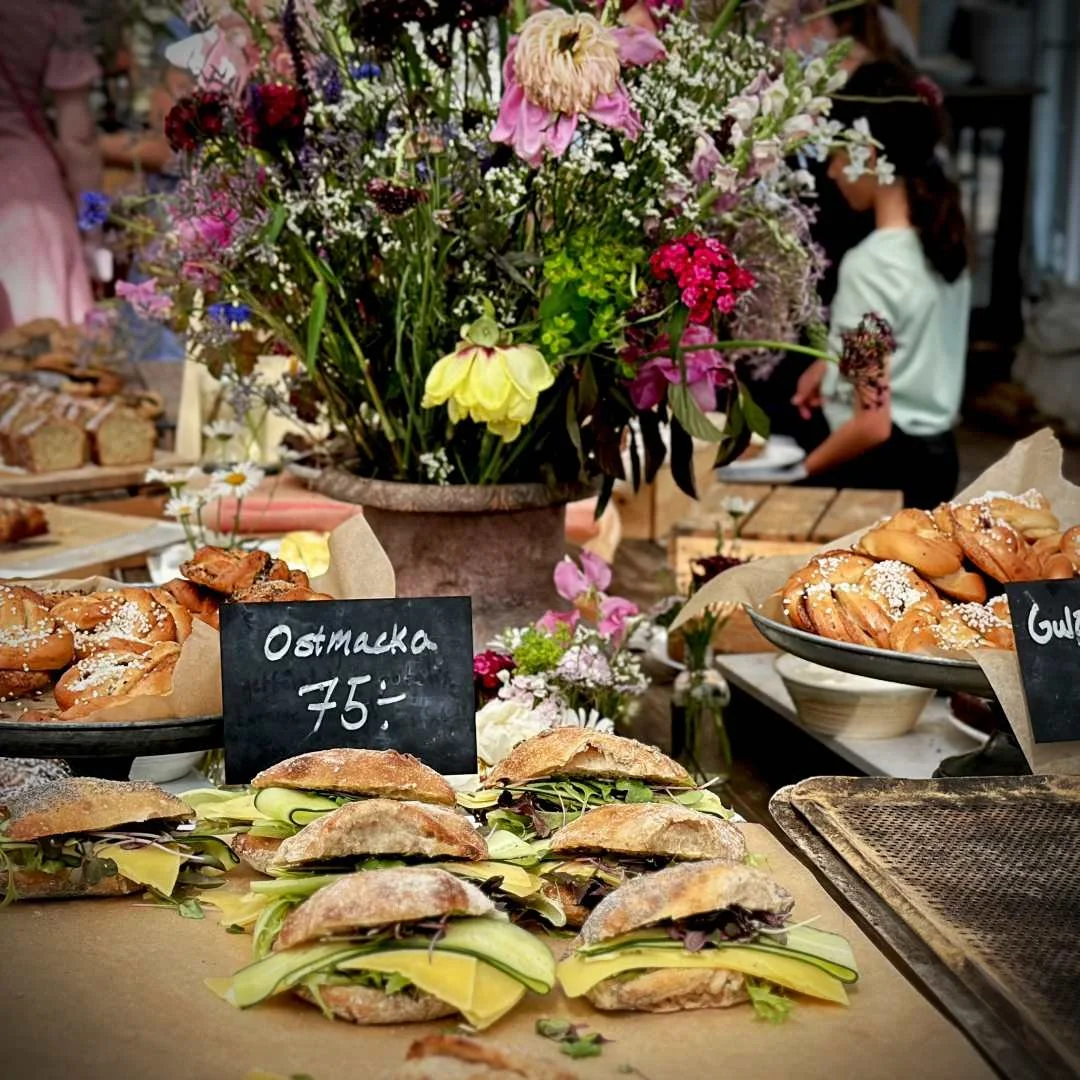 Several sandwiches filled with cheese, cucumber slices, and greens are arranged on baking paper in the foreground. Behind them, a small black chalkboard sign reads "Ostmacka 75