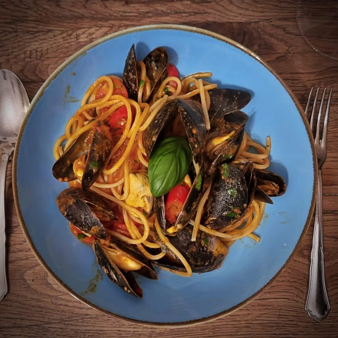 Spaghetti mit Miesmuscheln und Kirschtomaten in einer blauen Schüssel auf einem Holztisch im Restaurant Il Piccolo Principe in München.