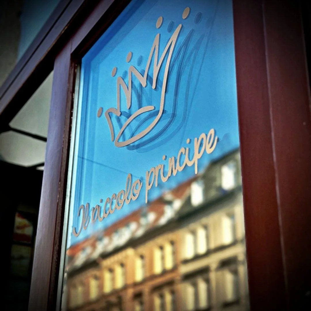 Close-up of a windowpane at restaurant Il Piccolo Principe featuring curved lettering and a crown against building reflections.
