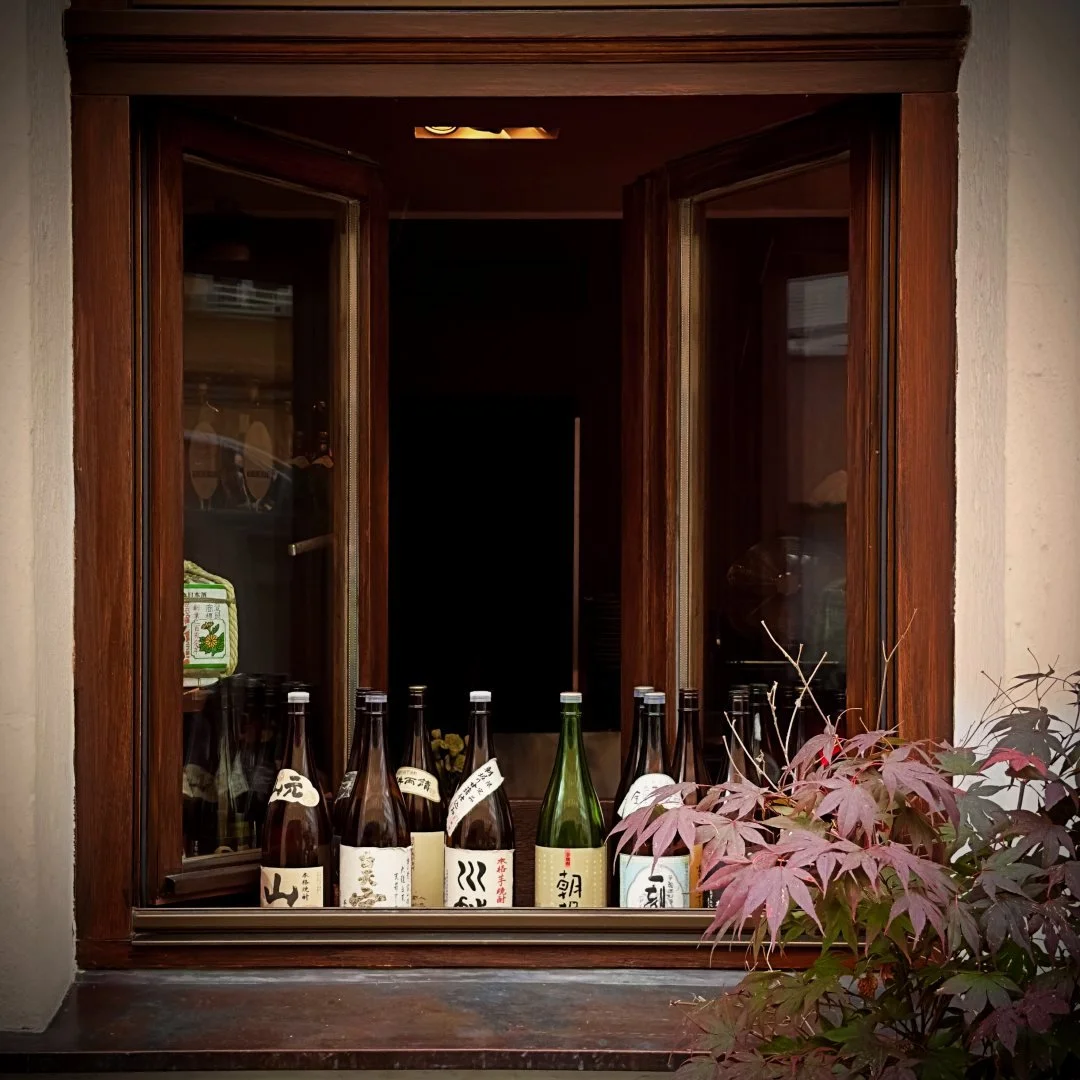 An open wooden window of the bar with sake and spirit bottles lined up on the windowsill beside red maple foliage in the foreground.