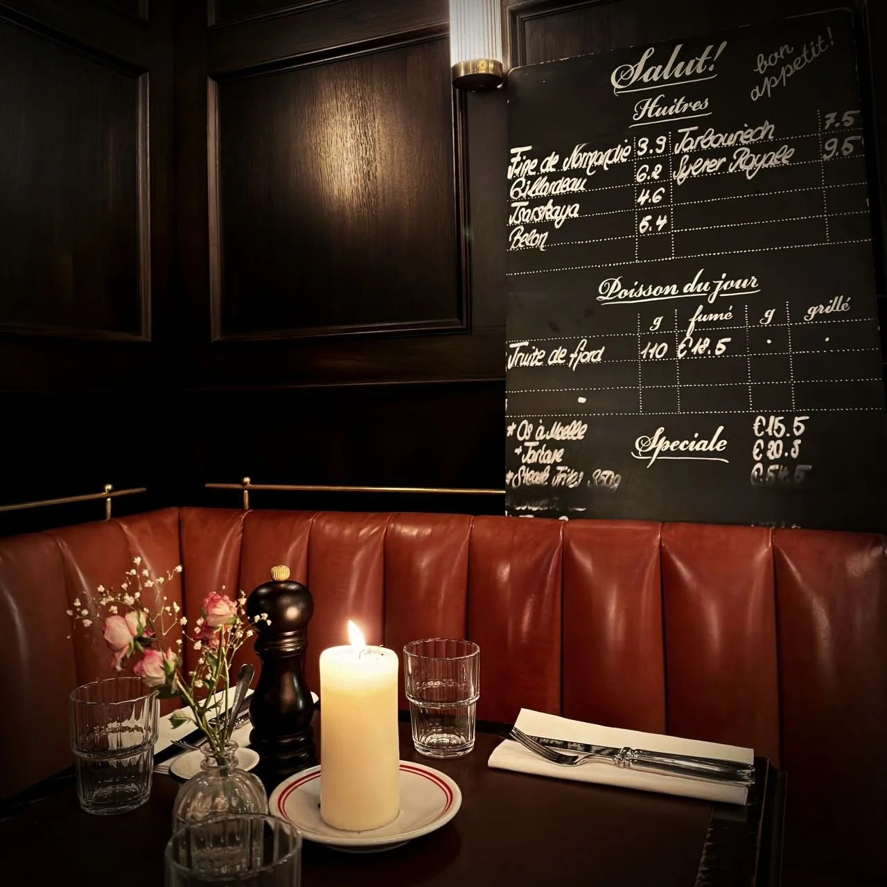 Set dining table with a burning candle, glasses, and flowers in front of a brown leather booth and a chalkboard menu at Petit Fritz.