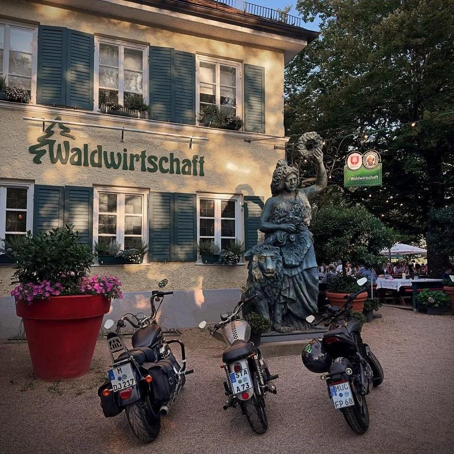 Exterior view of the entrance to Waldwirtschaft featuring a Bavaria statue with a lion, three parked motorcycles in front, and the beer garden in the background.