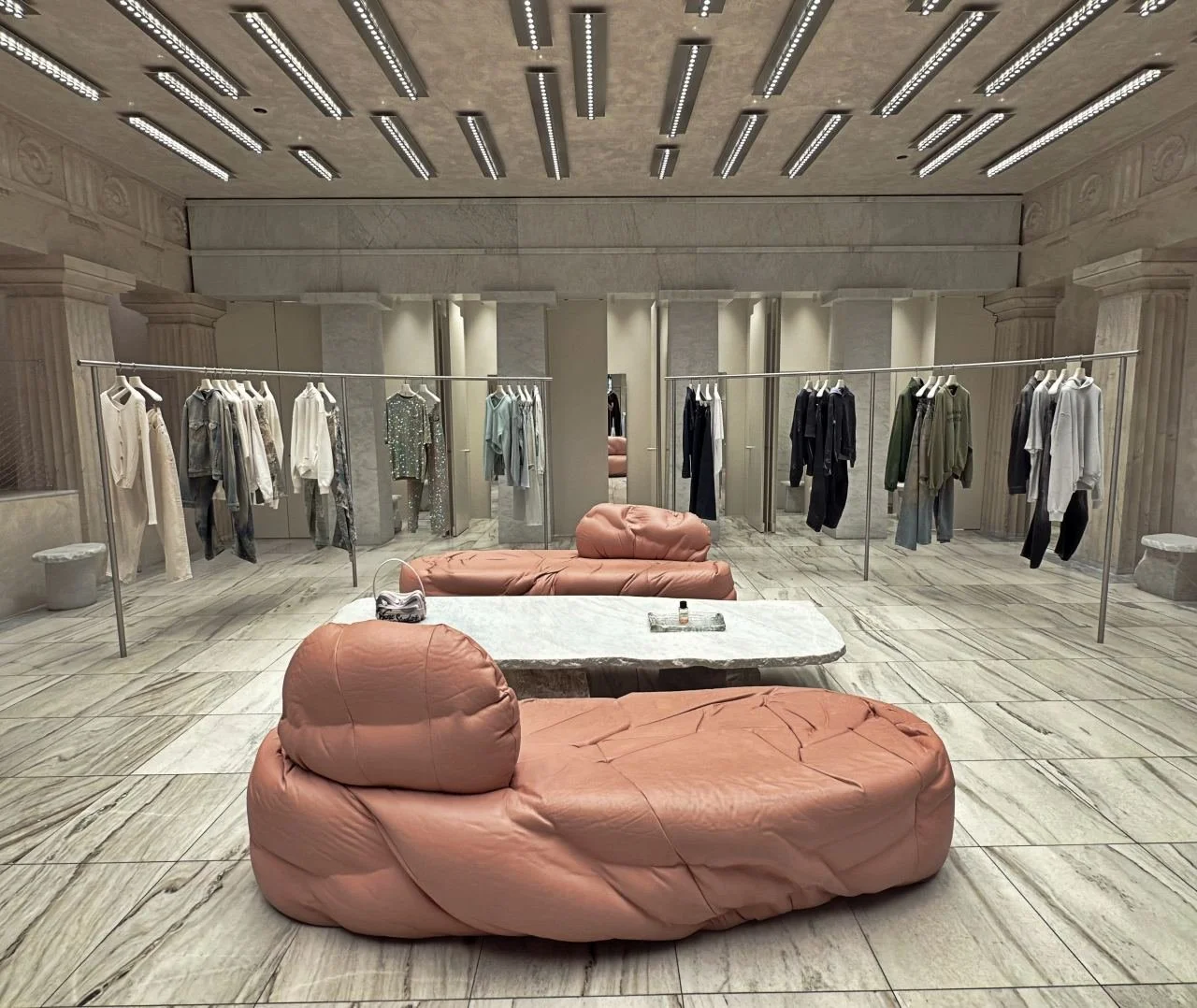 Acne Studios retail space in Stockholm featuring marble flooring, a symmetrical layout, clothing racks with minimalist fashion, and soft upholstered seating in muted pink tones.