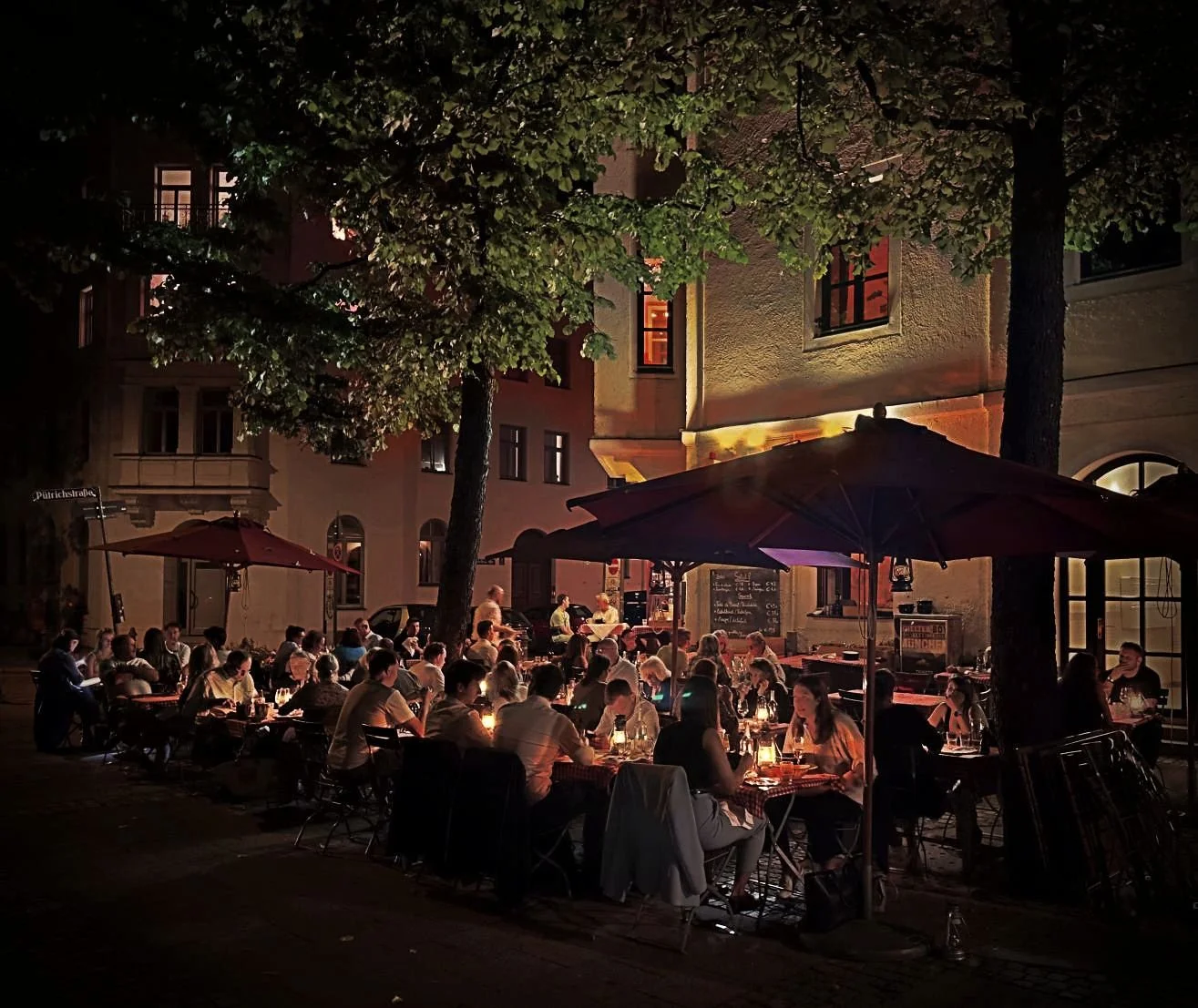 Busy outdoor terrace of restaurant Chez Fritz at night with guests dining under umbrellas and trees by candlelight.