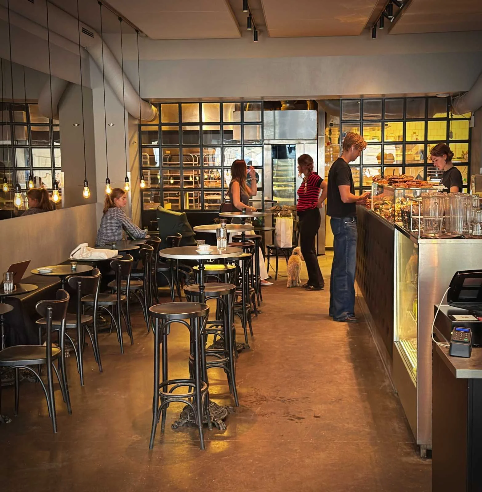 The interior of a modern cafe with bistro tables, bar stools, and hanging light bulbs. Customers line up at the long counter on the right, and a glass partition walls off the bakery area in the background.