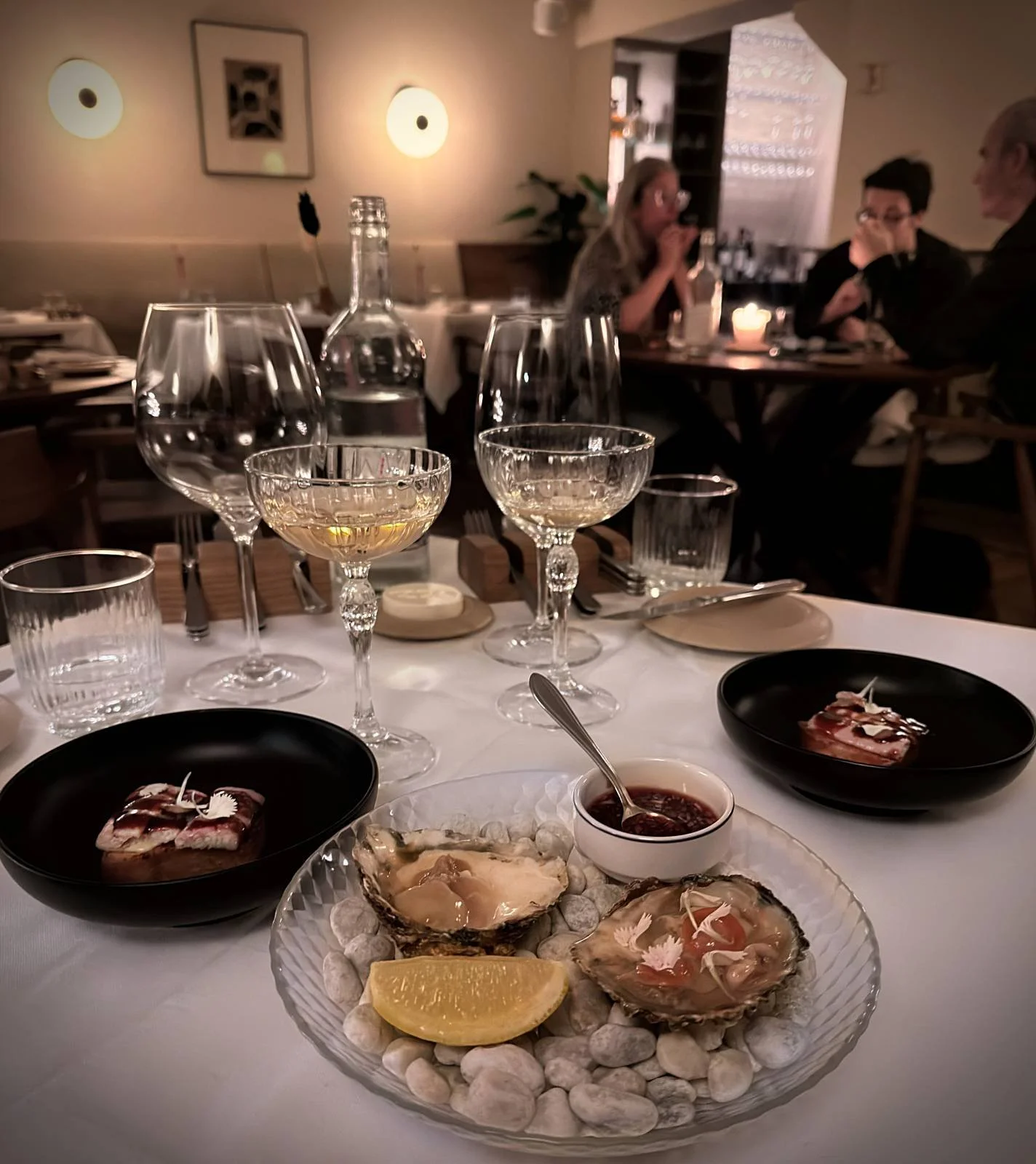 Ein gedeckter Tisch im Restaurant R21 in Amsterdam mit Austern auf Steinen, einer Zitronenspalte und zwei Vorspeisen in schwarzen Schalen. Im Hintergrund stehen Champagnergläser und Wasserflaschen.