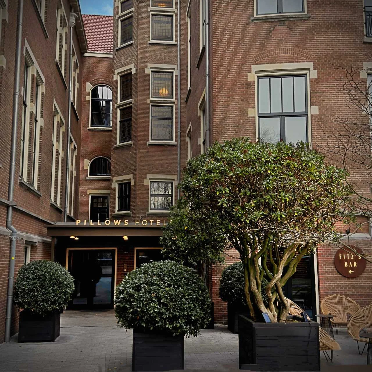 Die Backsteinfassade des Pillows Hotels zeigt dunkle Fensterrahmen und den Eingangsbereich mit großen Grünpflanzen in schwarzen Kübeln im Innenhof.