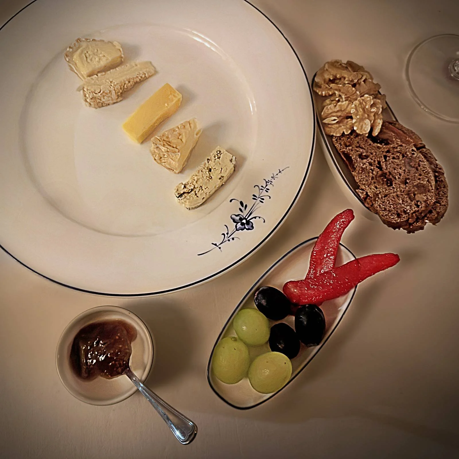 Eine Auswahl verschiedener Käsesorten auf einem weißen Teller mit blauem Muster. Dazu werden Brot, Walnüsse, Weintrauben und eine Beilage in kleinen Schalen im Restaurant R21 serviert.