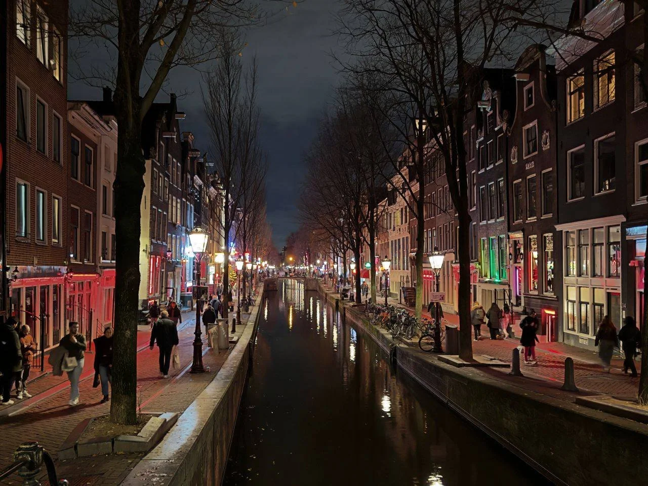 Beleuchtete Gracht im Amsterdamer Rotlichtviertel bei Nacht mit historischen Häusern, Spiegelungen im Wasser und Passanten.