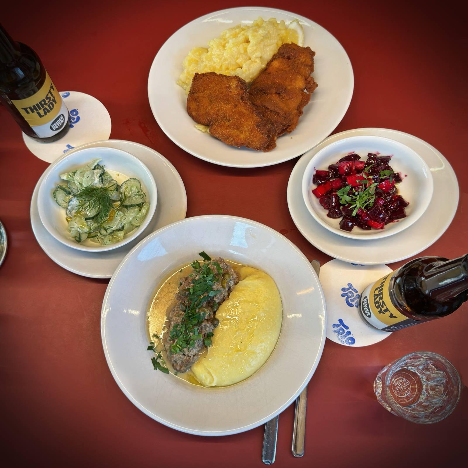 A plate with a rollmops, pickled cucumbers, a lemon wedge, and a pickled egg in a bowl on a red table. A plate with bread and butter and a bottle of beer stand behind it.