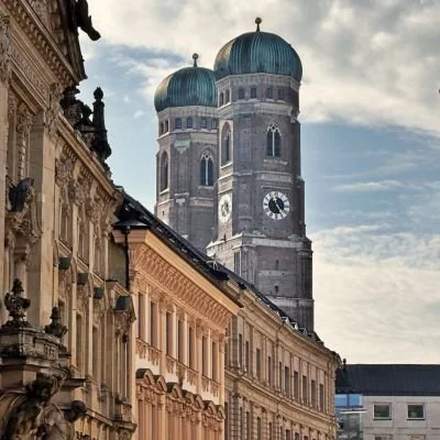 Die markanten Zwiebeltürme der Frauenkirche ragen über die prachtvollen Fassaden der Münchner Altstadt. Die Perspektive betont die historische Architektur und das traditionelle Herz der bayerischen Landeshauptstadt.
