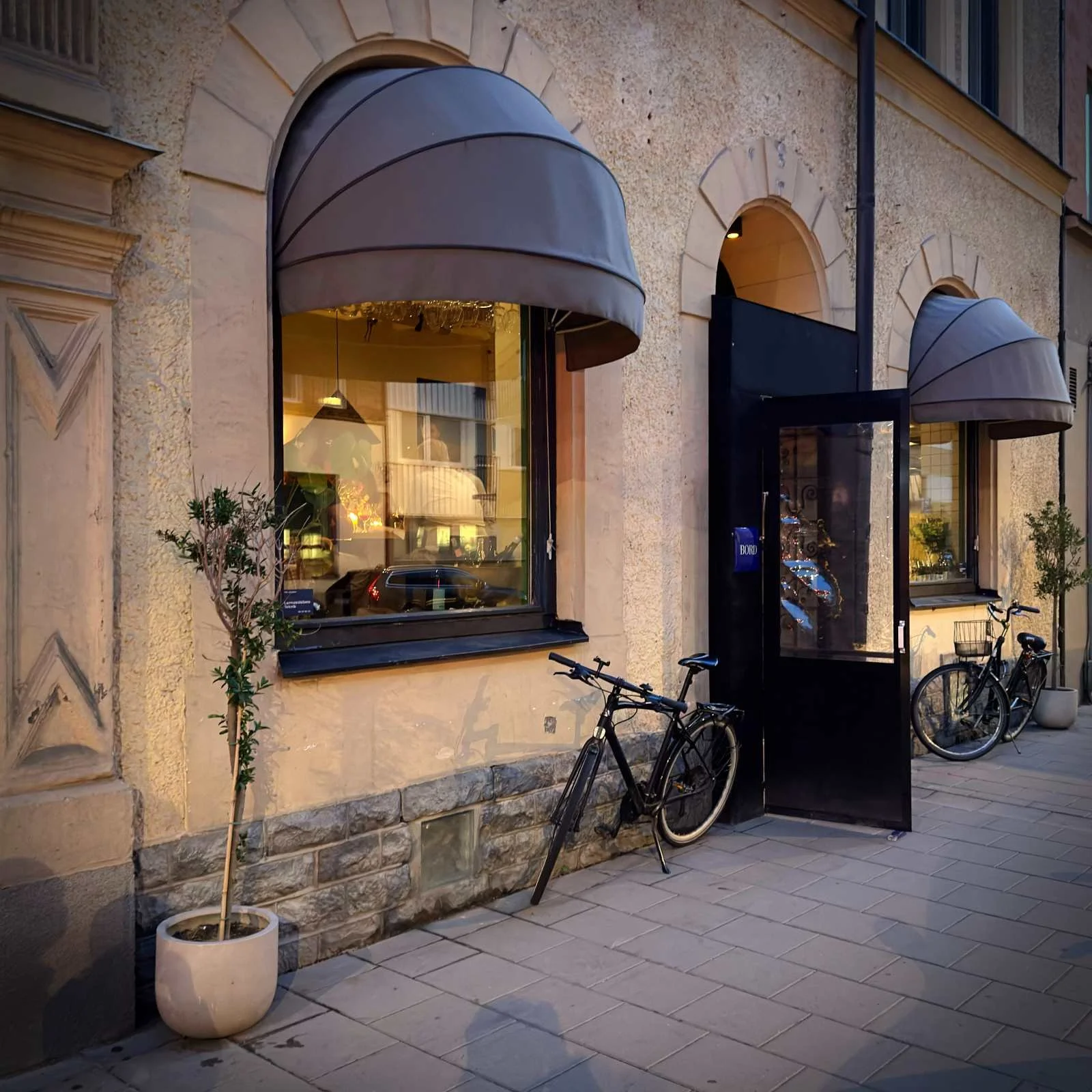 Die Außenansicht des Restaurants Bord mit hellbrauner Putzfassade, Rundbogenfenstern und grauen Markisen. Ein schwarzes Fahrrad steht neben der geöffneten Eingangstür, links befindet sich eine Topfpflanze auf dem Gehweg.