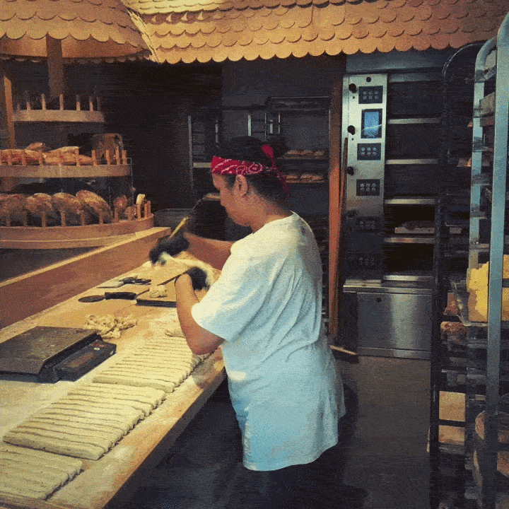 View through the window of Lillebrors Bageri showing a baker working with dough at a wooden table. The white lettering "Lillebrors Bageri" is visible on the window glass, with a baking oven in the background.