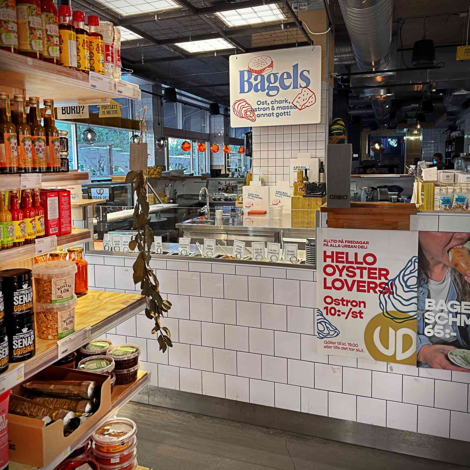 A white-tiled service counter inside Urban Deli featuring signs for bagels and oysters. Grocery shelves stocked with sauces, mustard, and fried onions are visible on the left.