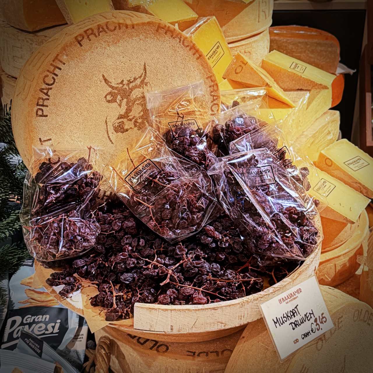 m Shop De Kaaskamer in Amsterdam liegen Muskat-Trauben in einem Korb vor großen Käselaiben. Ein Preisschild zeigt den Preis für Muskaat Druiven an, im Hintergrund stapelt sich gelber Käse.
