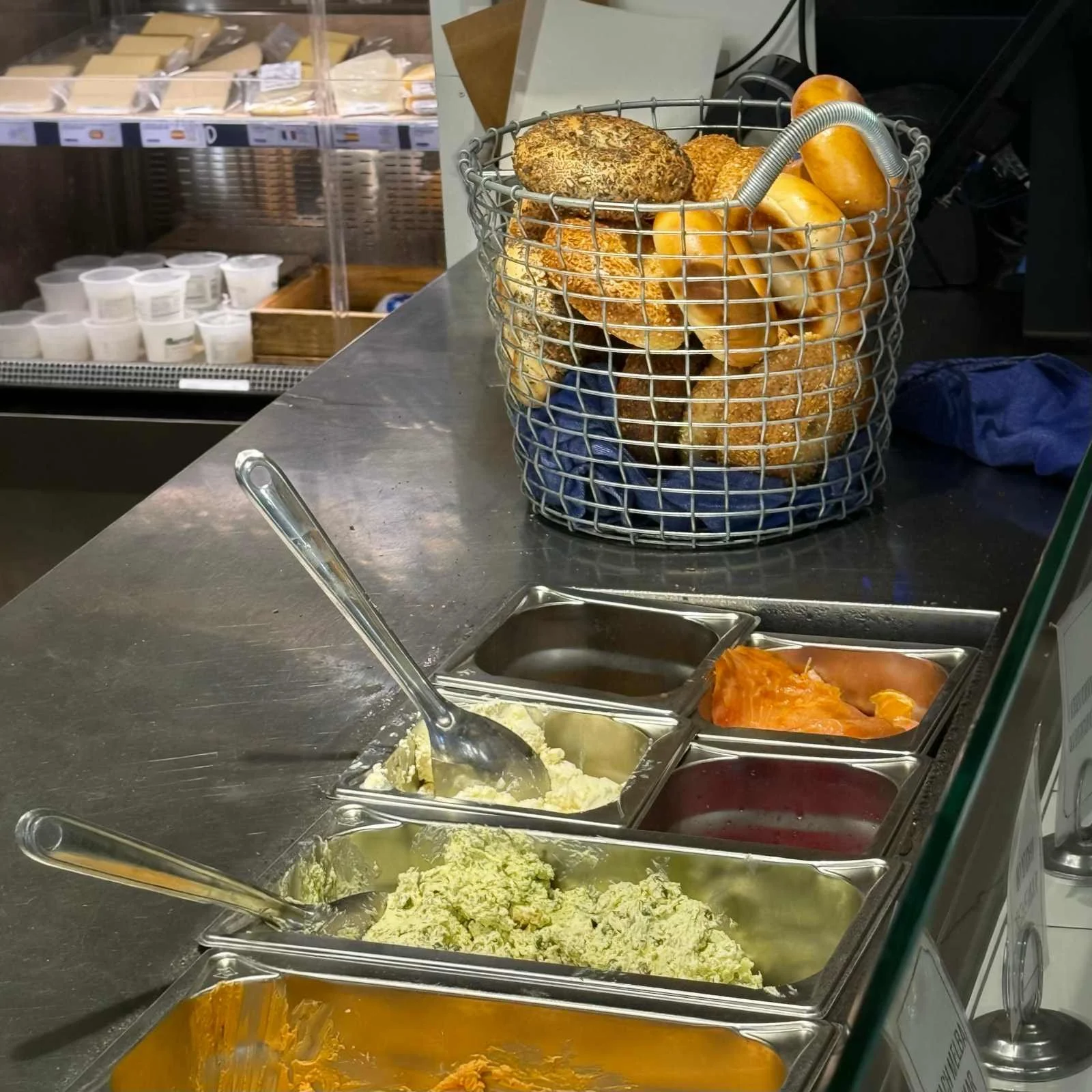 A stainless steel counter featuring food containers filled with various cream cheese spreads and smoked salmon. A wire basket filled with fresh, assortment bagels stands behind them.