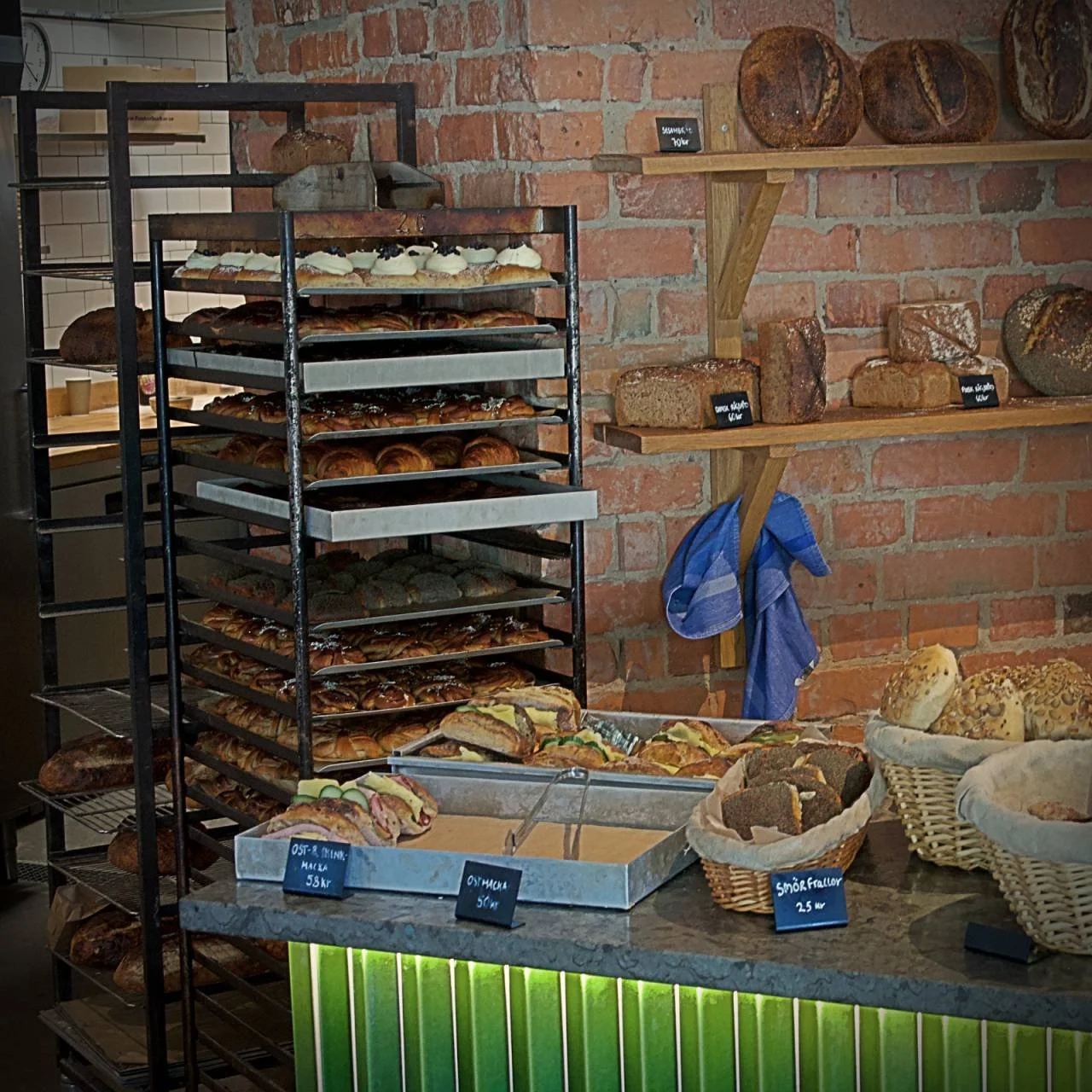 A tall rack trolley filled with Semlor and cinnamon buns stands in the open bakery in front of the brick wall. Trays with sandwiches and baskets of rolls lie on the counter in front.