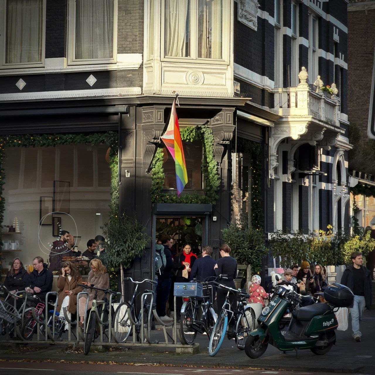 Guests sit outside „The Coffee District“ cafe near Vondelpark. A rainbow flag hangs above the entrance. Many bicycles and a green electric scooter are parked on the sidewalk in front.