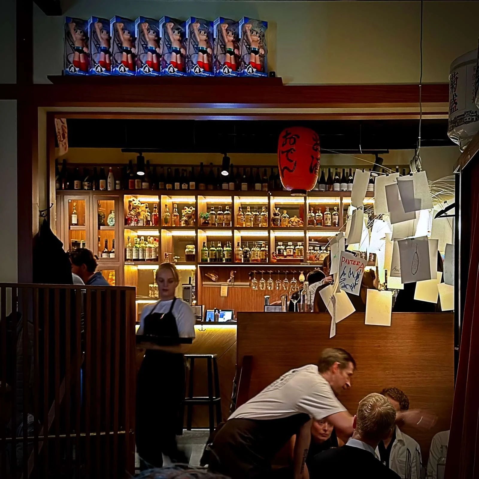 View of the bar at Miyakodori restaurant in Stockholm featuring backlit bottle shelves, staff, and Astro Boy figures on a shelf above.
