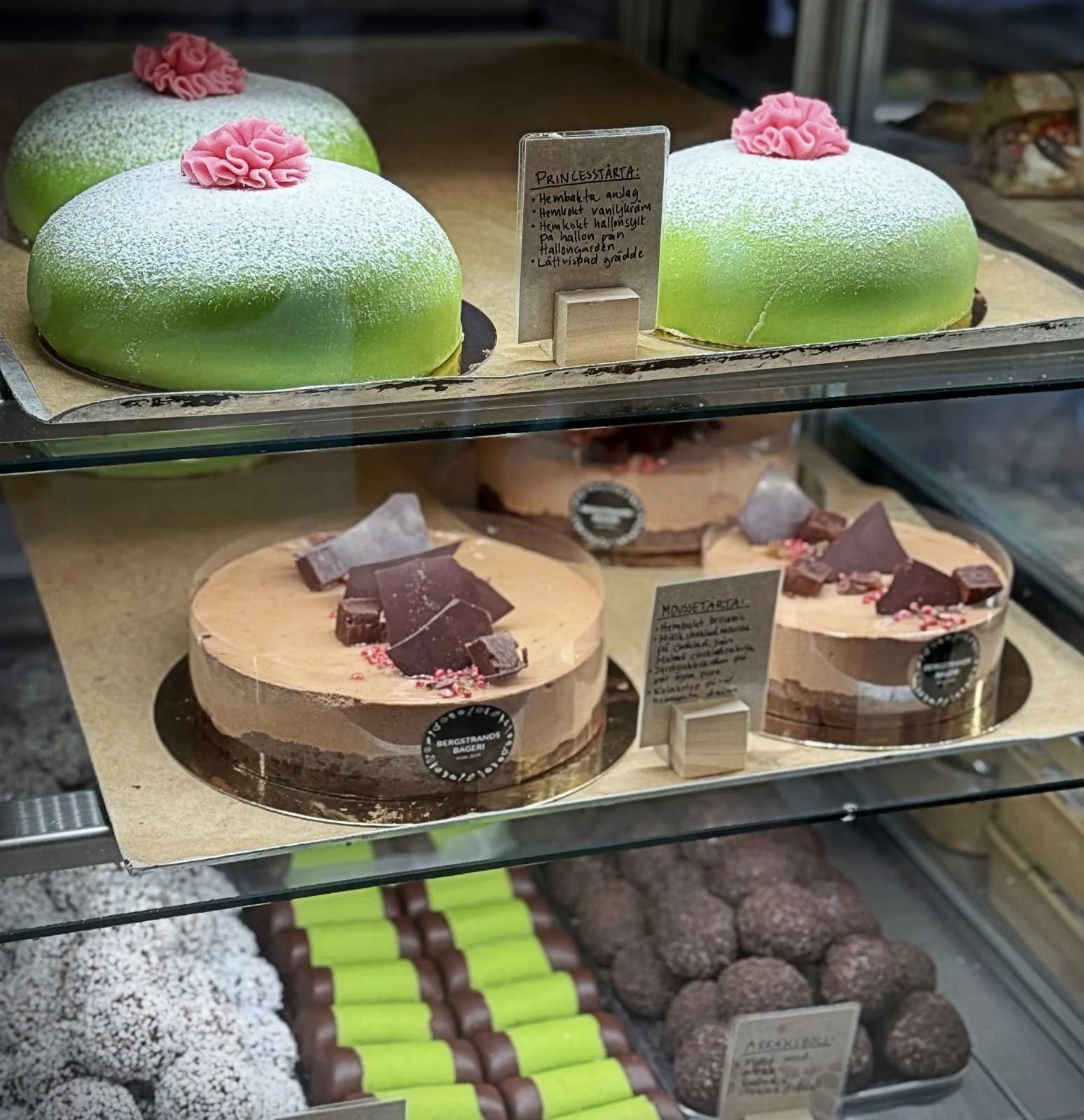 Eine gläserne Kuchenvitrine in der Bergstrands Bageri in Stockholm, gefüllt mit verschiedenen Torten, darunter eine grüne Prinzessinnentorte (Prinsesstårta) und Schokoladenkuchen.