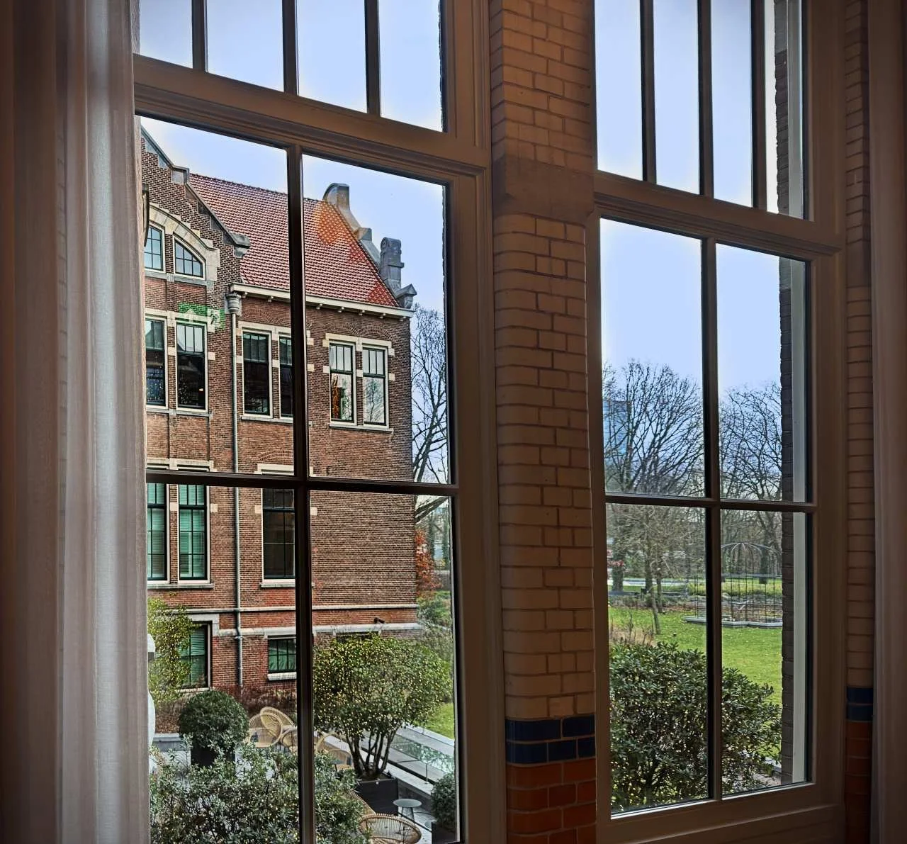 Blick durch hohe Sprossenfenster auf die benachbarte Backsteinfassade und den herbstlichen Oosterpark im Pillows Hotel Amsterdam.