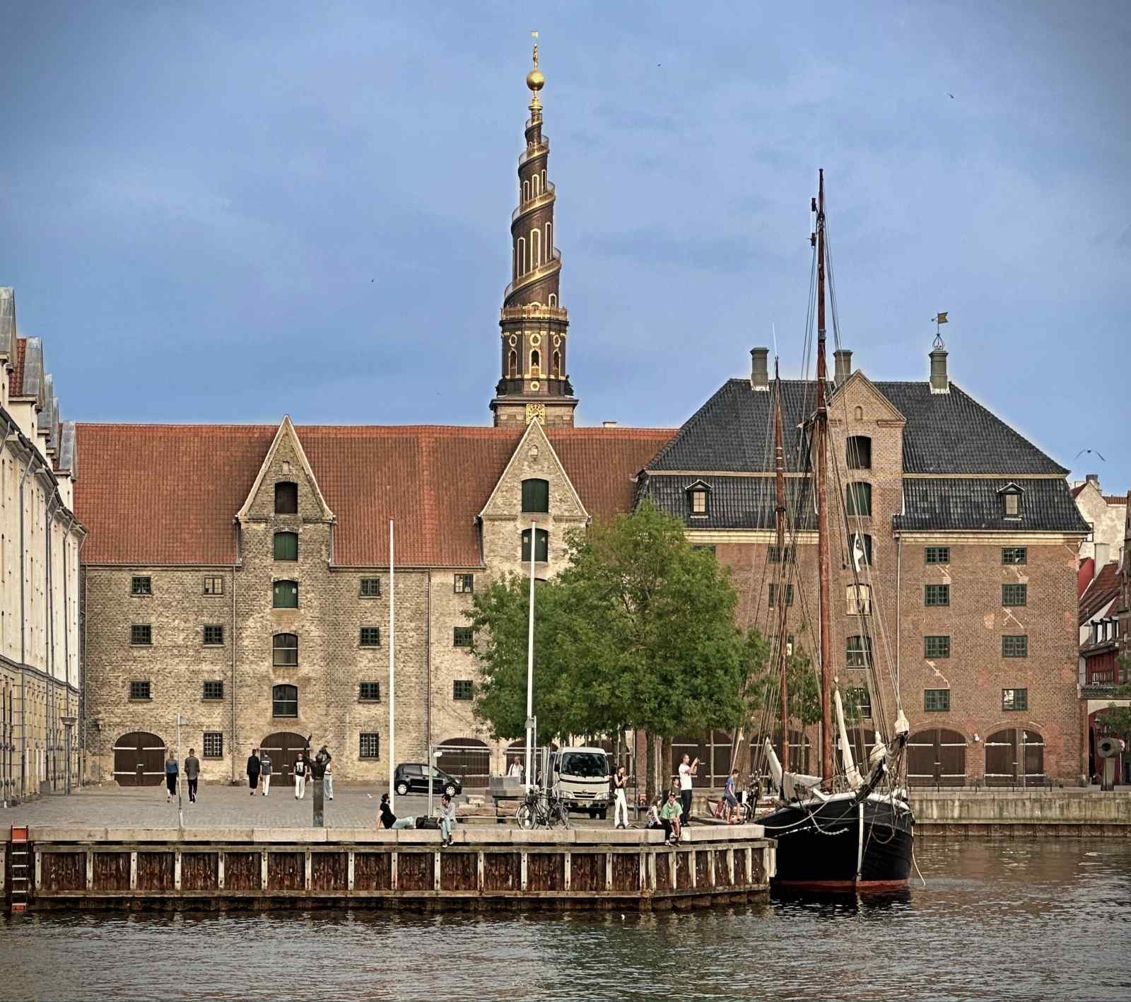 Ein Blick auf ein Hafenbecken im Stadtteil Christiania in Kopenhagen. Am Ufer stehen Bäume und mehrere Holzhäuser, im Wasser spiegelt sich die Vegetation.