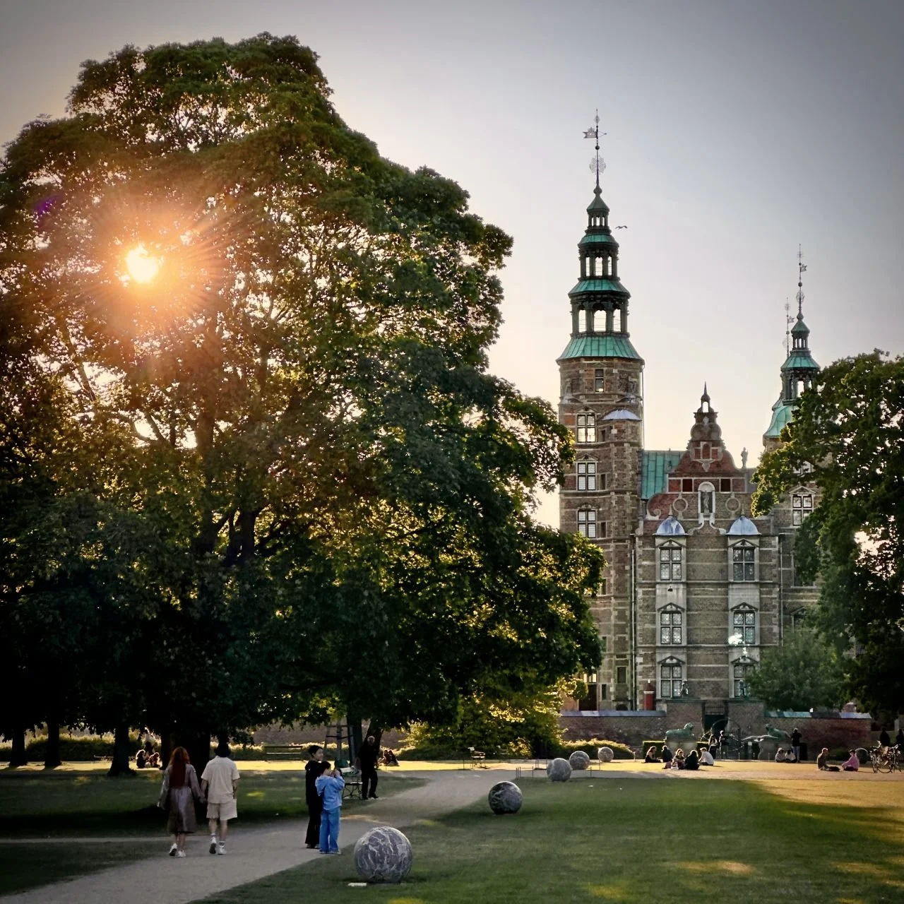 View through Kongens Have in Copenhagen towards Rosenborg Castle with brick towers and copper roofs. Lawn with footpath, trees, stone sculptures and people relaxing in the park.