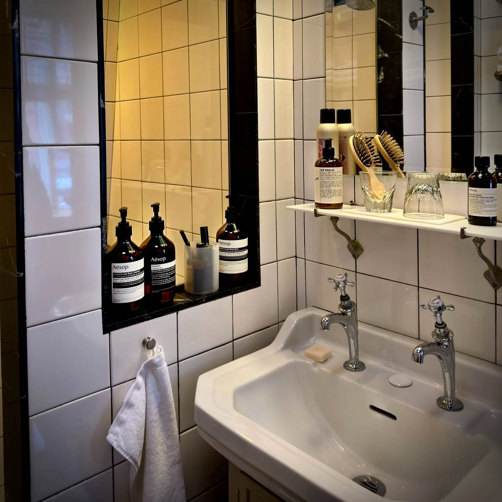 Drei braune Spenderflaschen der Marke Aesop stehen in einer Halterung an einer weißen Fliesenwand im Badezimmer. Daneben befindet sich eine gläserne Duschwand mit silbernem Rahmen.