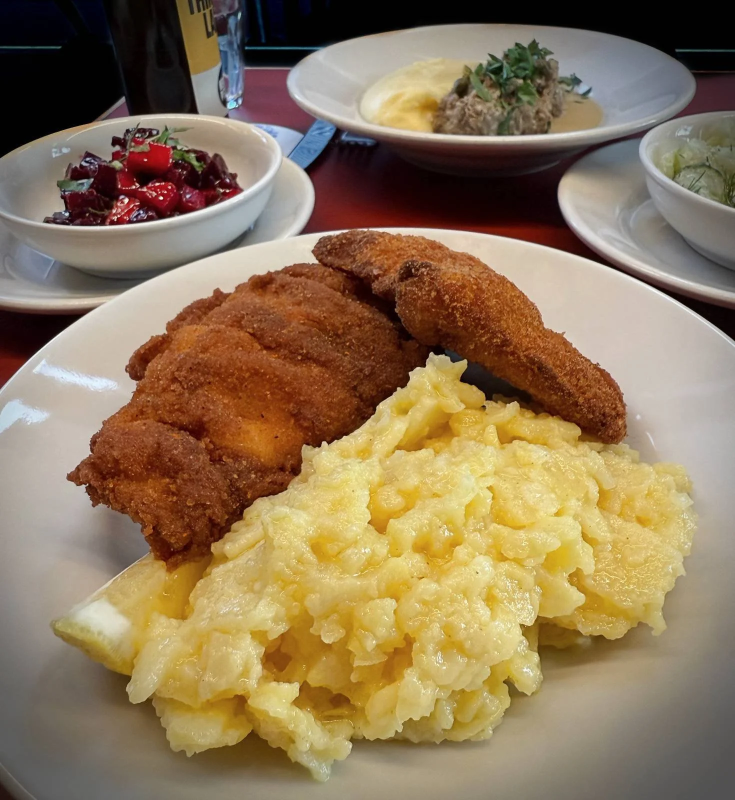 Close-up of a plate with breaded fried chicken and potato salad on a red table. Bowls with beetroot salad, cucumber salad, and meatballs stand in the background.