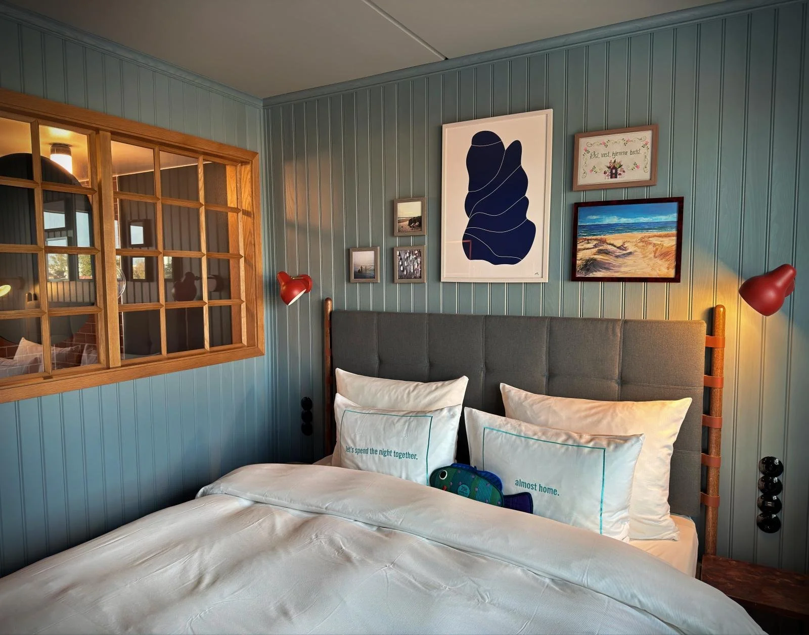 Frisch gemachtes Doppelbett mit weißer Bettwäsche und Zierkissen vor türkisfarbenen Wandpaneelen im 25hours Hotel Kopenhagen. Bilderrahmen und rote Wandlampen hängen über dem grauen Kopfteil.