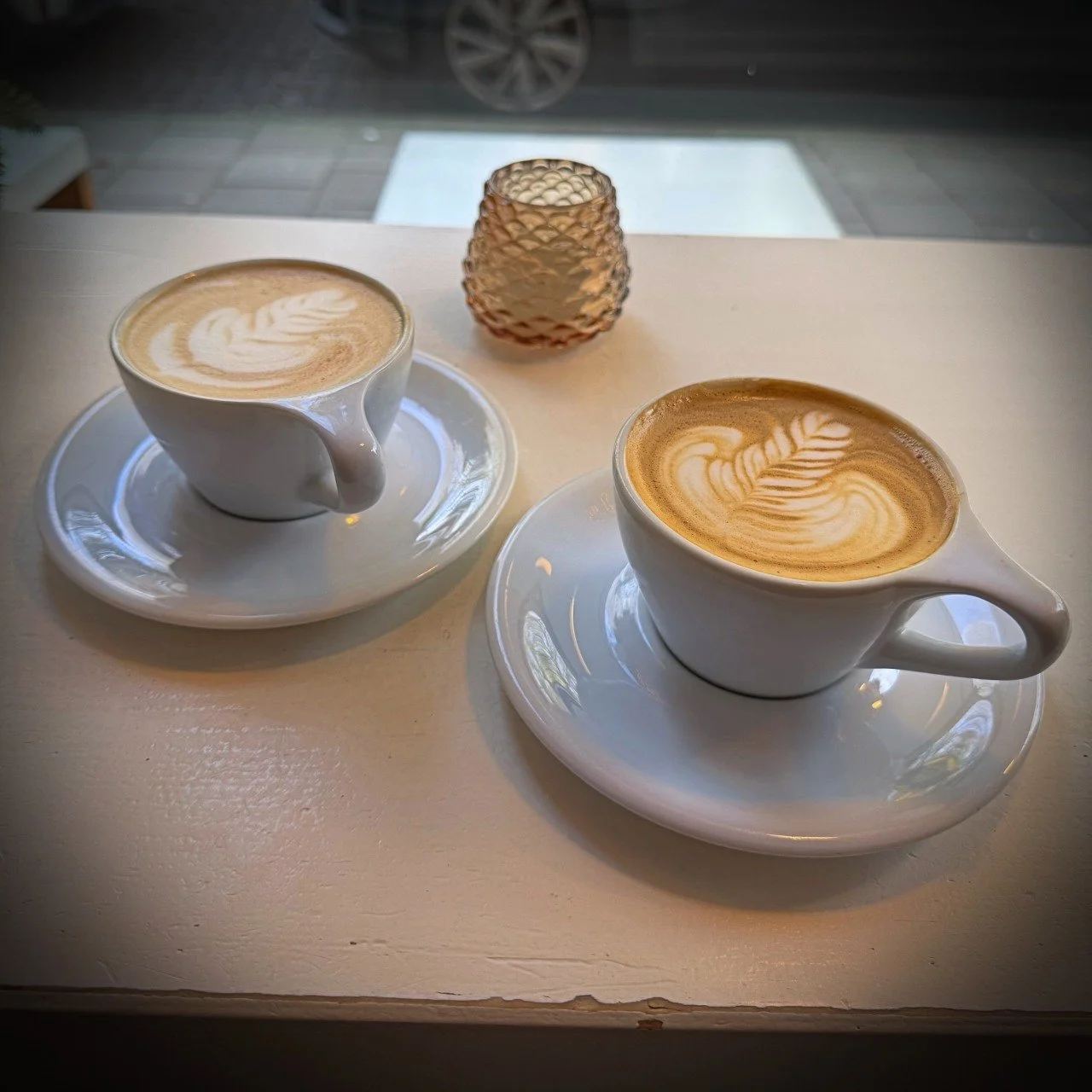 Two cappuccinos with latte art in white cups on saucers placed on a light table by the window inside Scandinavian Embassy café in Amsterdam, with a small glass candle holder.