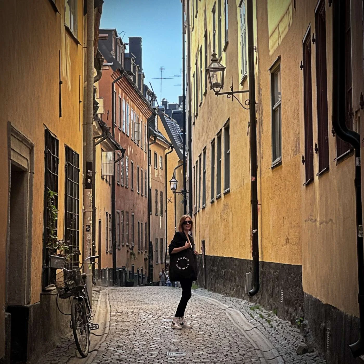 ine enge Gasse mit Kopfsteinpflaster führt durch Gamla Stan, die Altstadt von Stockholm. Hohe, ockerfarbene Fassaden säumen den Weg, auf dem eine Person steht.