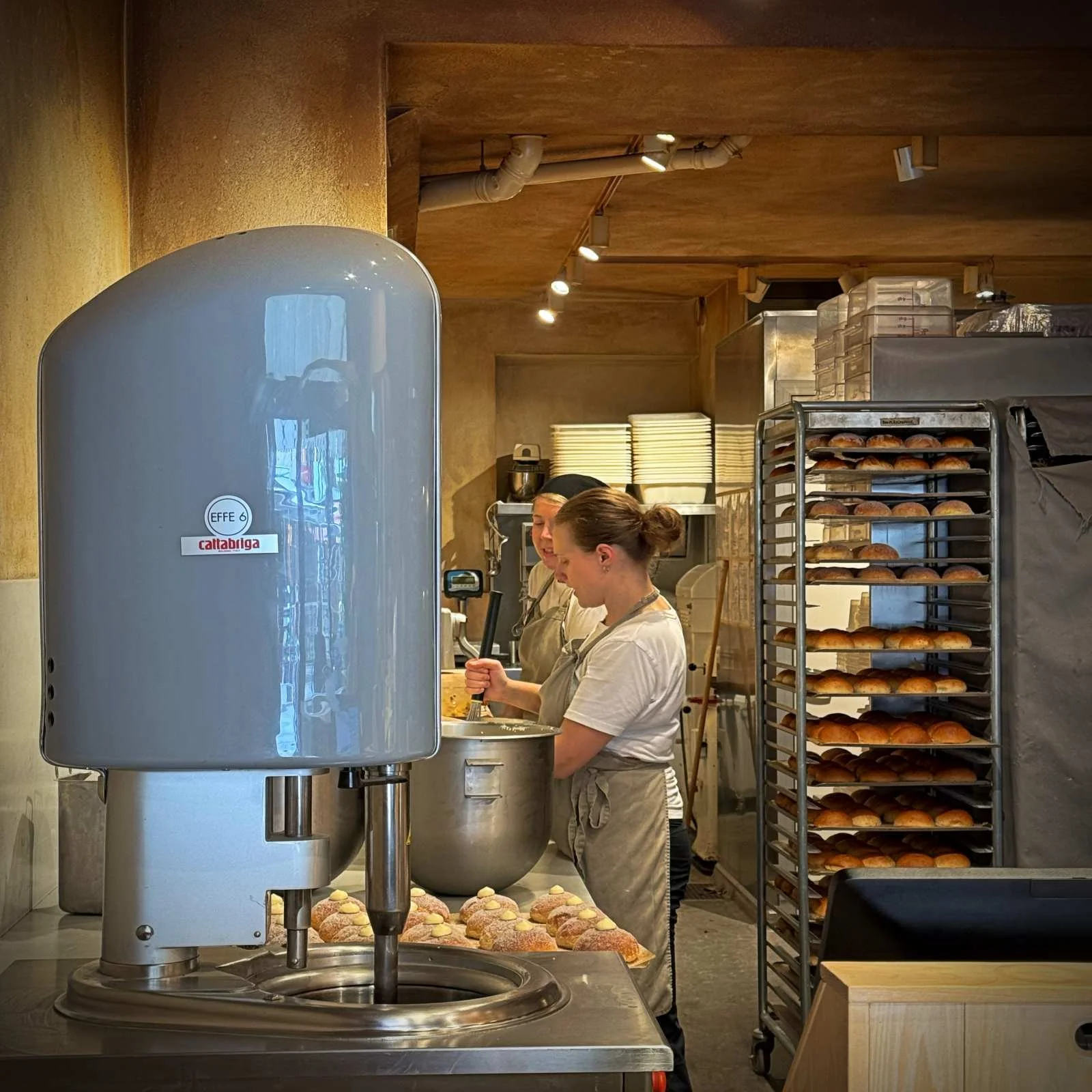 Blick in eine Backstube mit einer grauen Cattabriga-Eismaschine im Vordergrund. Zwei Bäckerinnen arbeiten an einem großen Edelstahltopf, daneben stehen gefüllte Gebäckstücke und ein Stufenwagen voller frischer Brötchen.