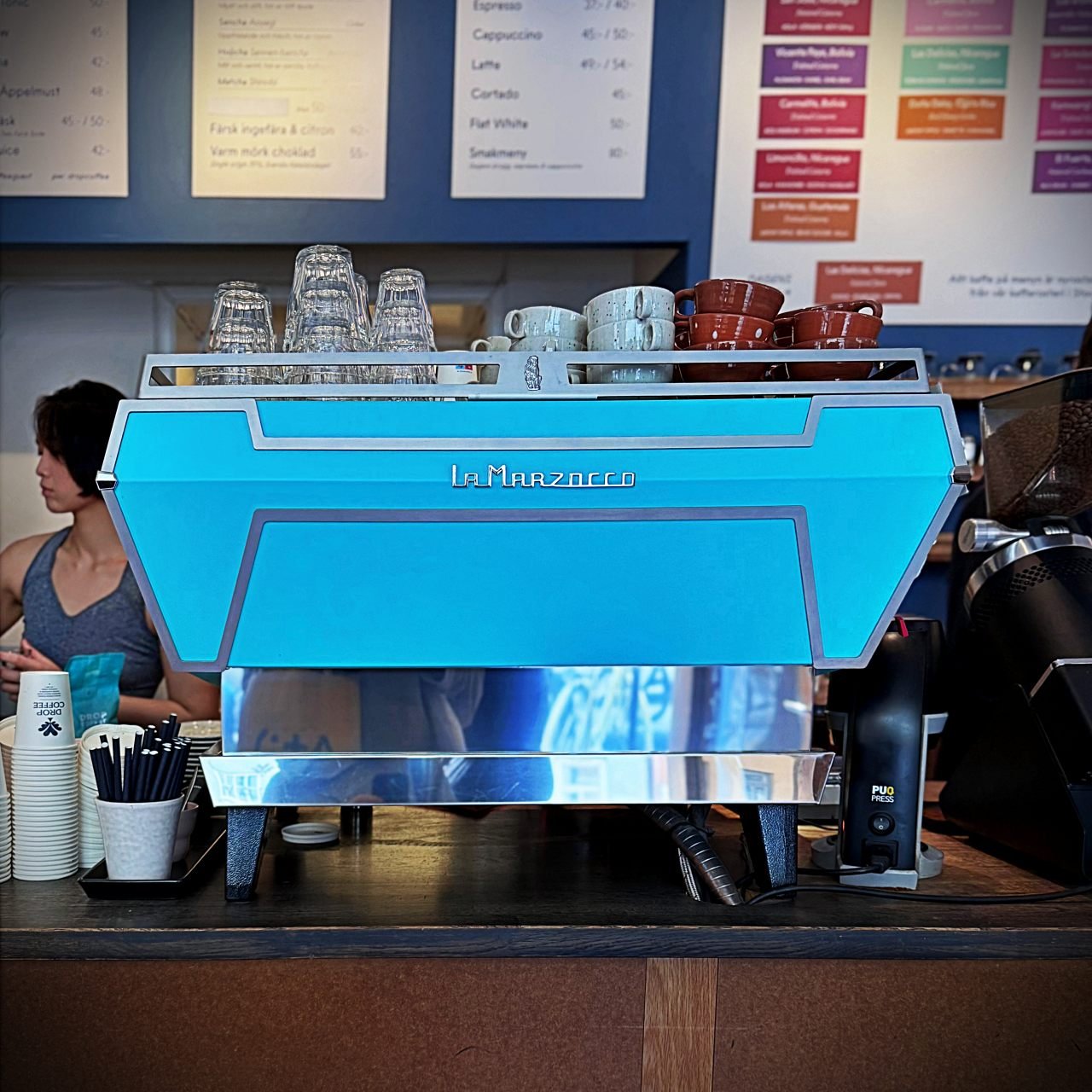 Eine weiße Kaffeemaschine der Marke La Marzocco steht auf dem Holztresen des Cafés Drop Coffee in Stockholm. Im Hintergrund bedient eine Person im blauen Oberteil die Kaffeemühle, daneben hängen Kaffeetassen an der Wand.