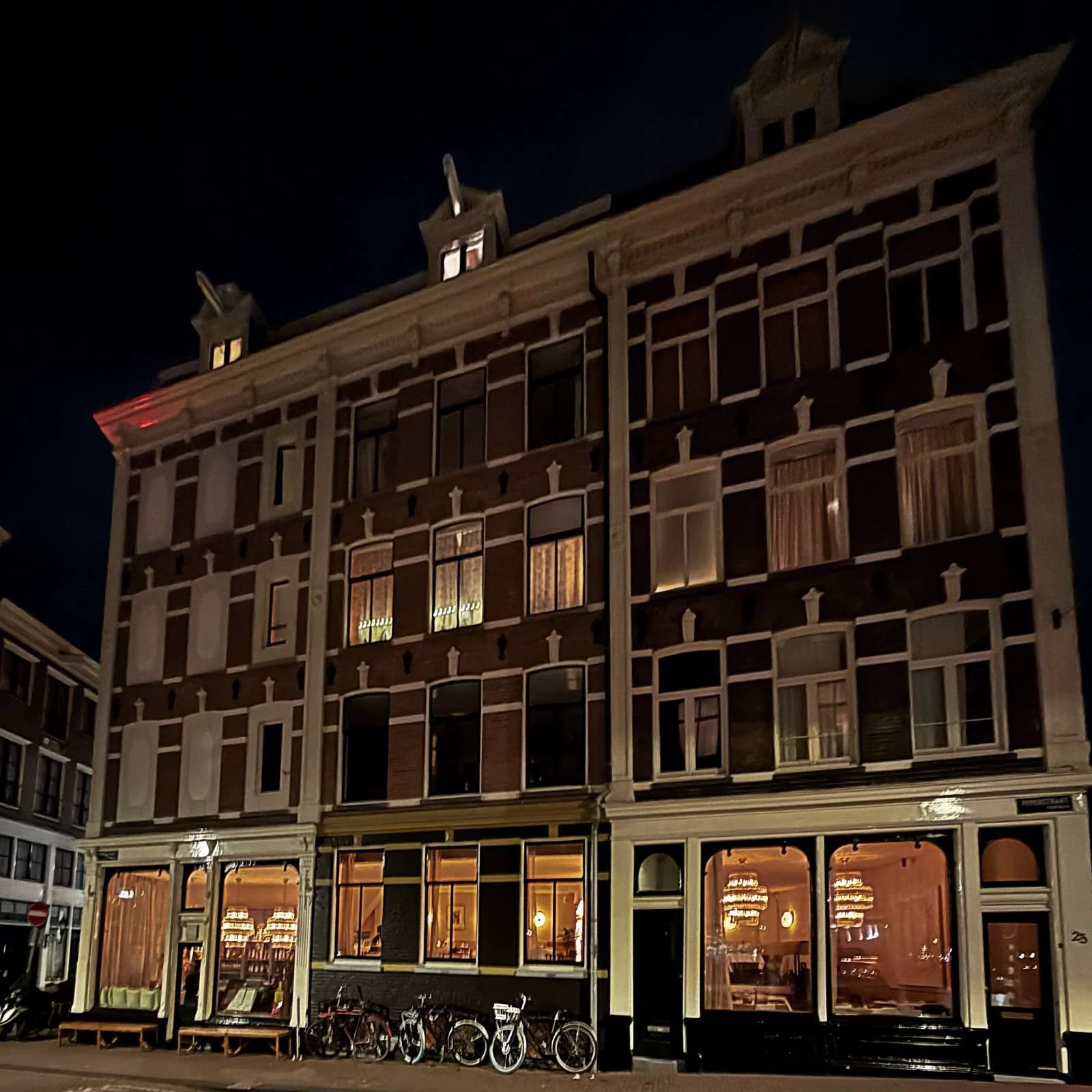 Eine nächtliche Aufnahme der Backsteinfassade des Restaurants R21 in Amsterdam. Mehrere beleuchtete Fensterfronten zeigen den Innenraum. Davor stehen einige Fahrräder auf dem Gehweg am Straßenrand.