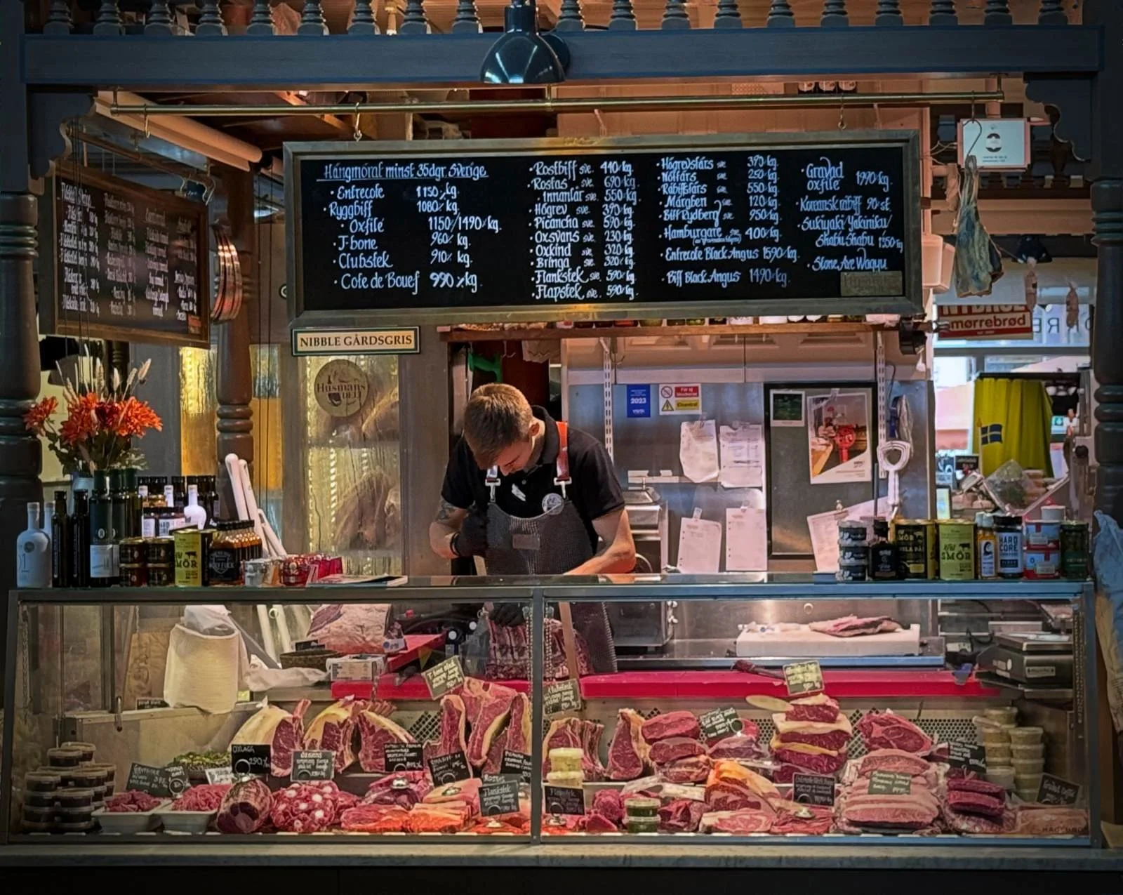 A staff member works behind the meat counter in Östermalms Saluhall. Various cuts of beef, such as Entrecôte and Oxfilé, are displayed in the glass case, with a large chalkboard showing prices hanging above.
