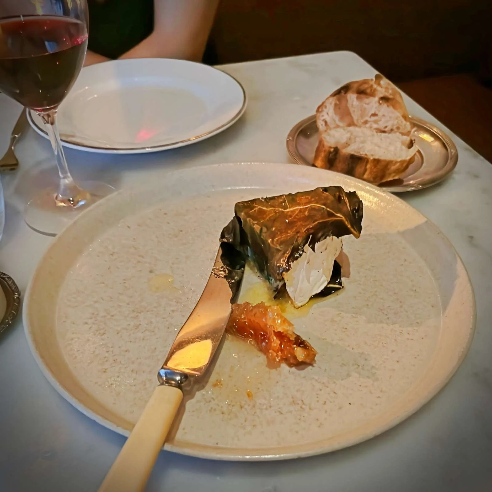 Ein Stück Käse in einem Feigenblatt serviert mit ein Stück Honigwabe auf einem beigefarbenen Teller. Im Hintergrund stehen ein Glas Rotwein und Brotscheiben auf einem Nebenteller.
