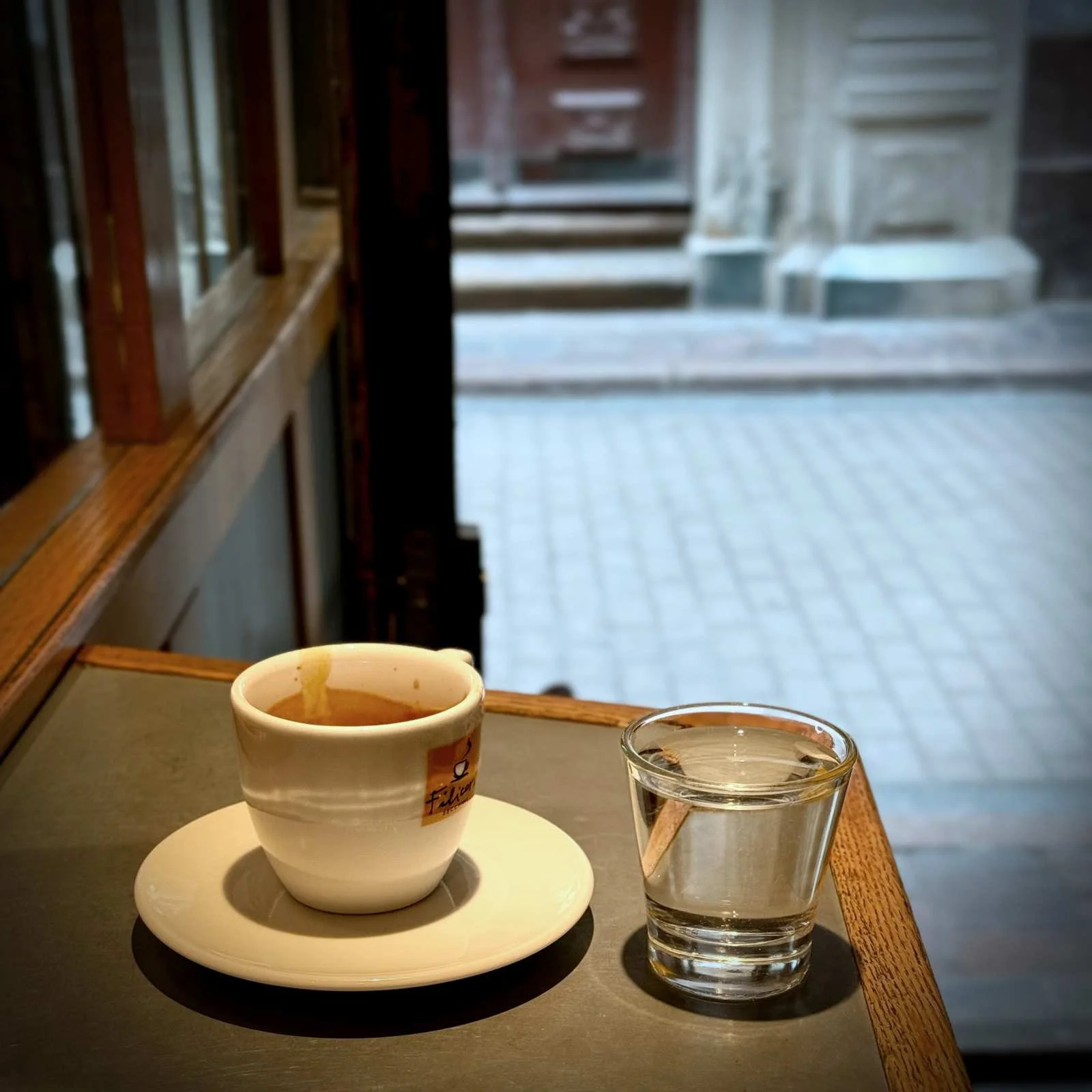 Eine weiße Tasse Espresso auf einer Untertasse neben einem kleinen Glas Wasser auf einem Tisch am Fenster.