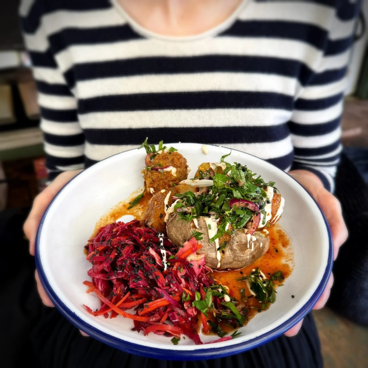 Gabi in a striped sweater holds a plate with a stuffed baked potato, fresh herbs, and colorful cabbage salad at Caspar Plautz.