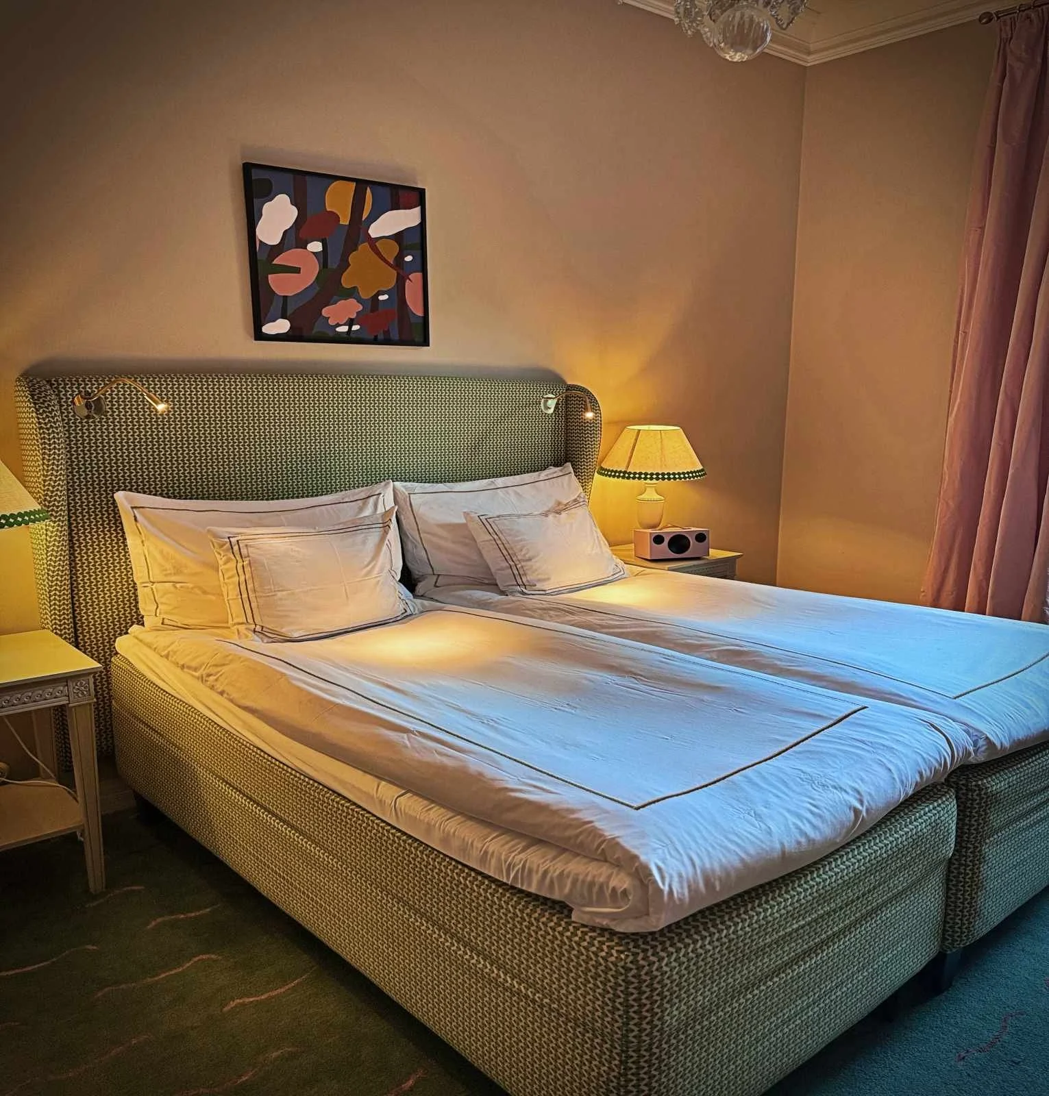Ein Hotelzimmer mit einem Doppelbett, das ein grün-weiß gemustertes Kopfteil aufweist. Das Bett ist weiß bezogen. Links und rechts stehen Nachttische mit Lampen.