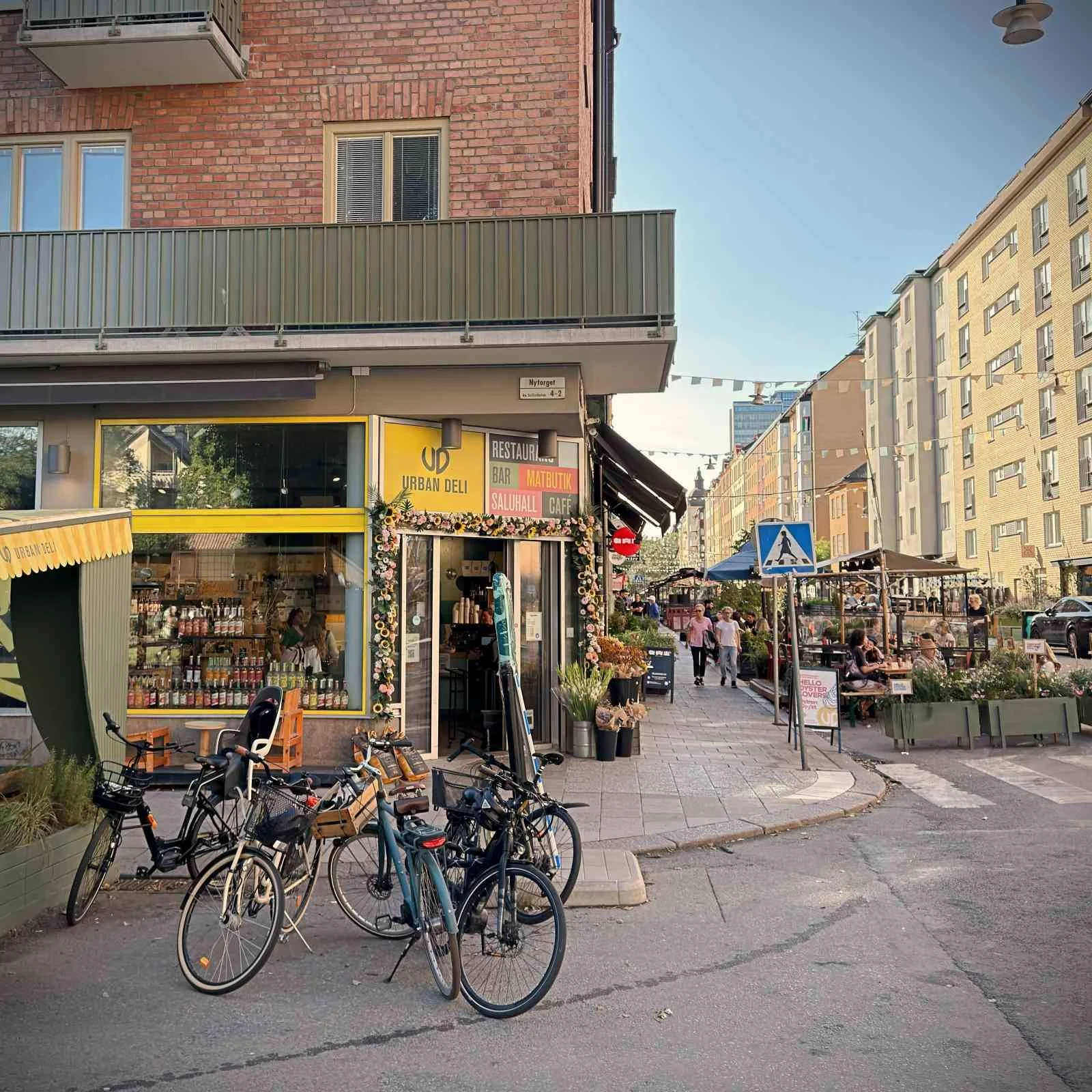 Exterior view of Urban Deli at Nytorget on a street corner of a brick building. Several bicycles are parked in front of the yellow-framed entrance, with a sidewalk featuring outdoor dining and pedestrians extending to the right.