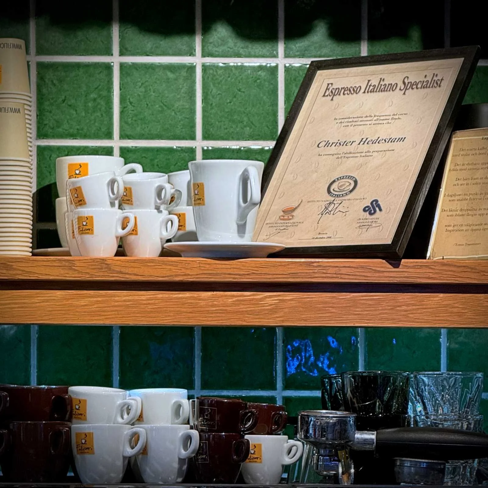 Weiß-braune Espressotassen auf Holzregalen neben einem gerahmten Zertifikat „Espresso Italiano Specialist“.