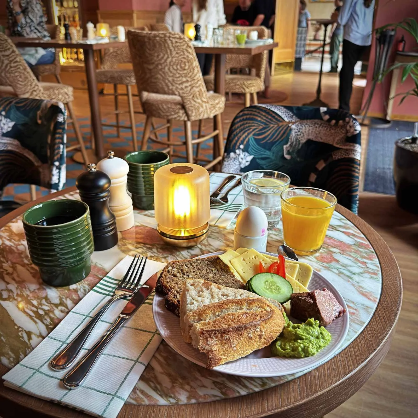 Ein runder Tisch mit Marmoroberfläche im Innenbereich von The Sparrow Hotel. Auf dem Tisch stehen ein Teller mit Brot, Käse, Gurkenscheiben und Avocado-Mousse, ein gekochtes Ei im Becher, ein Glas Orangensaft, ein Glas Wasser sowie Kaffeetassen.