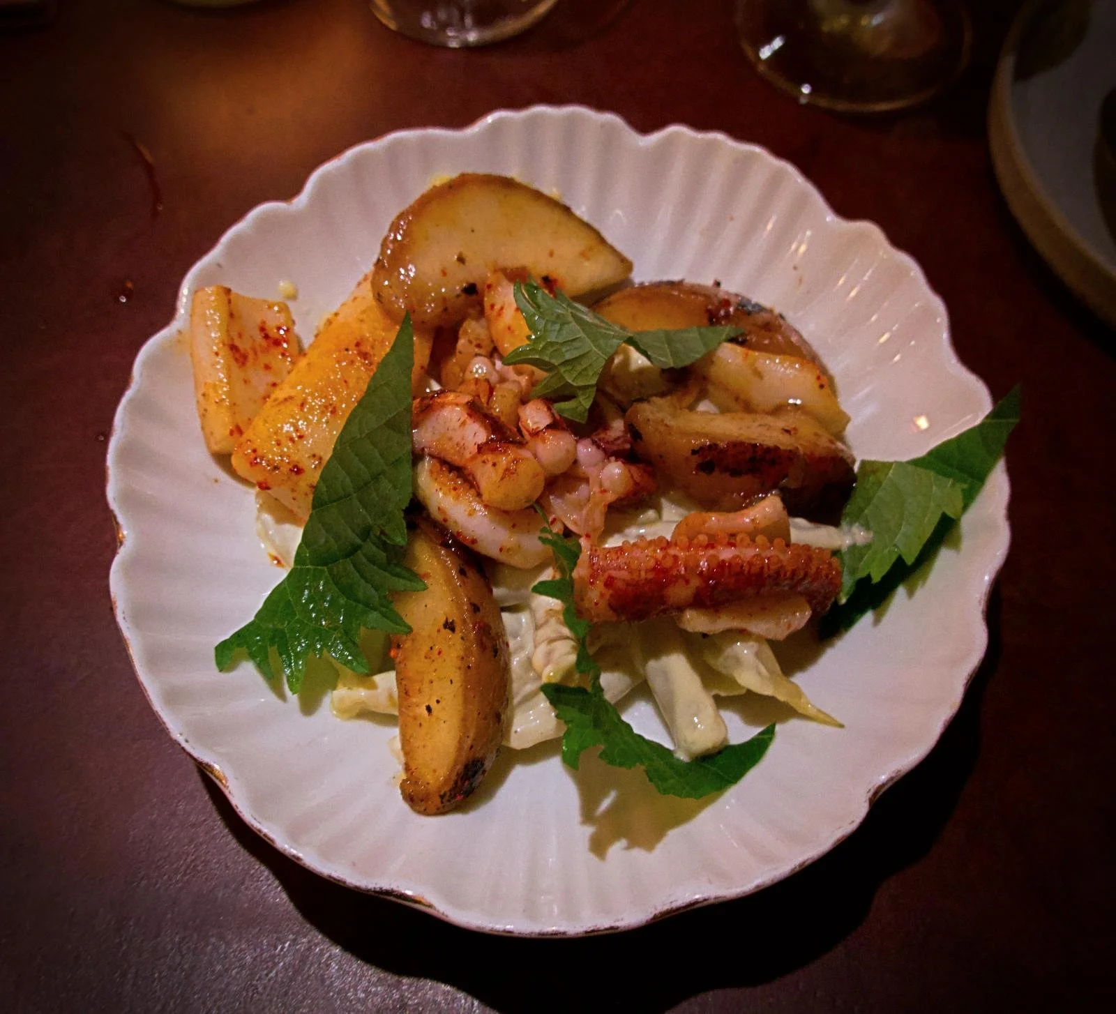 weiße Porzellanschale mit Wellenrand, in der gegrillter Tintenfisch, Pfirsichspalten, Bohnen und Shiso-Blätter angerichtet sind. Das Gericht wird auf einer dunklen Tischoberfläche im Restaurant Främmat präsentiert.
