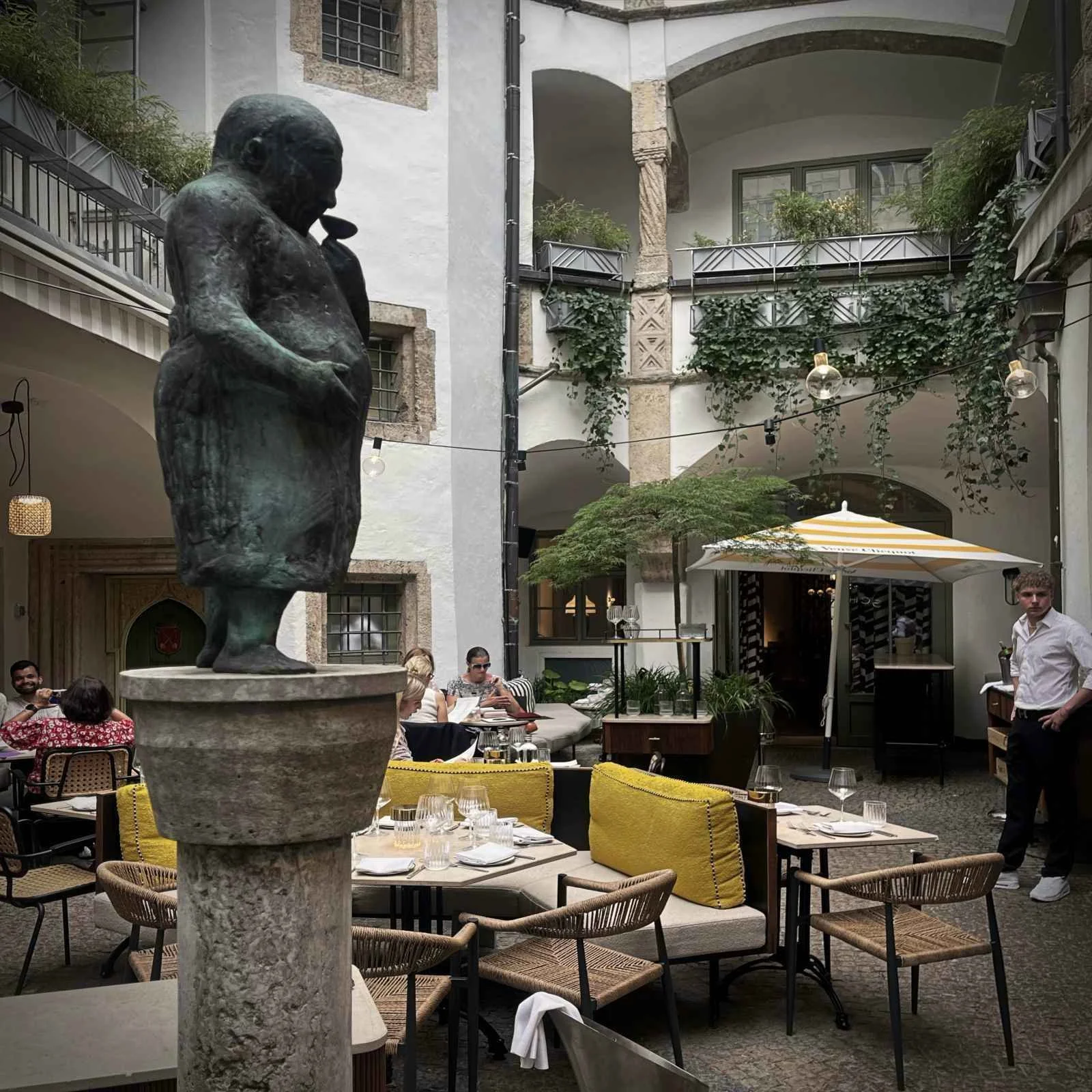 The planted Renaissance courtyard of restaurant Schreiberei in Munich featuring dining tables, yellow cushions, and a bronze sculpture in the foreground.