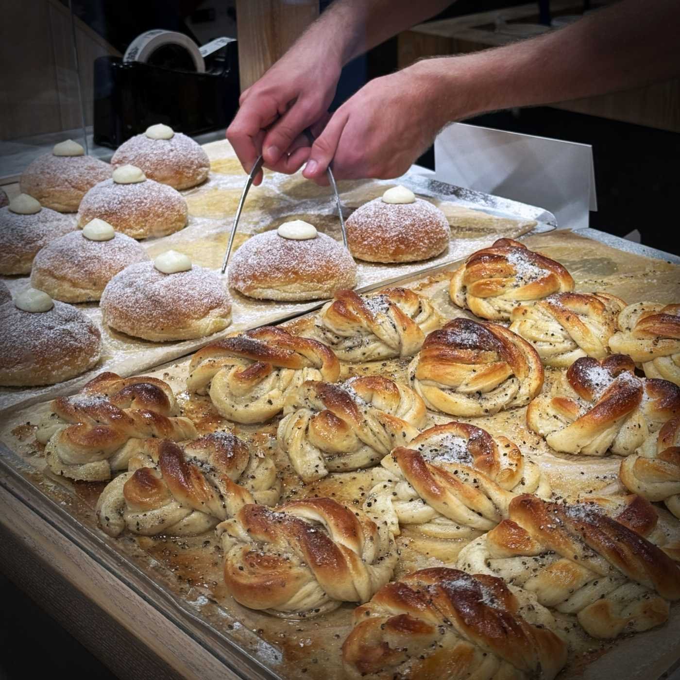 Zwei Backbleche mit frischem Gebäck. Im Vordergrund liegen Kardamomknoten (Kardemummabullar), dahinter stehen mit Puderzucker bestreute und Creme gefüllte Brötchen (Krämbullar), die von zwei Händen mit einer Zange gegriffen werden.